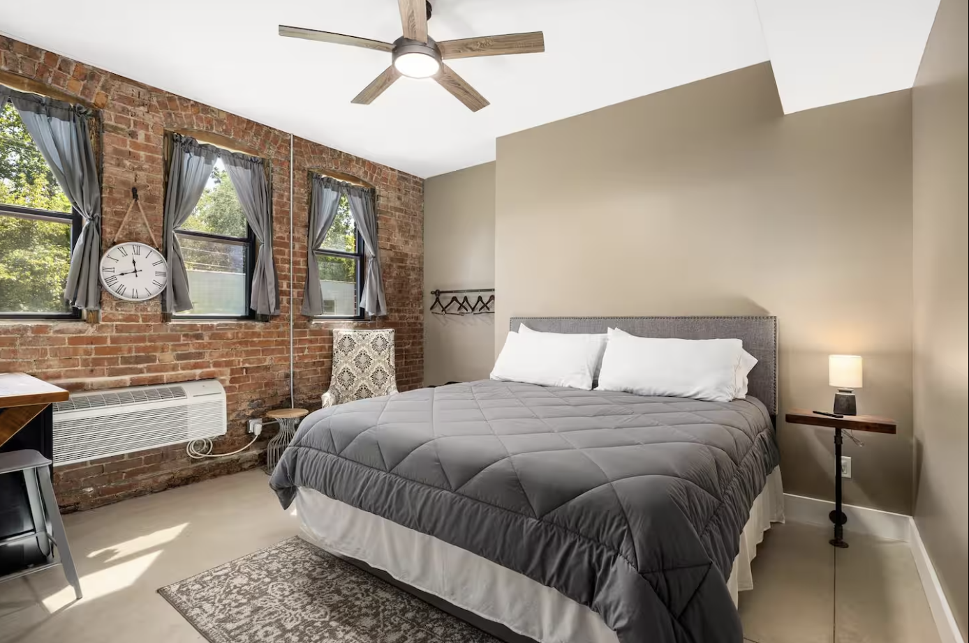A bedroom with a brick wall, three windows with grey curtains, a ceiling fan, a bed with grey bedding, a small side table with a lamp, and a chair near the windows.
