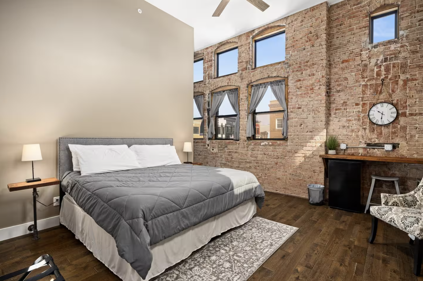 Bedroom with exposed brick wall, large windows, gray bed with white pillows, two bedside lamps, a clock, a small desk with a plant, and a patterned chair.