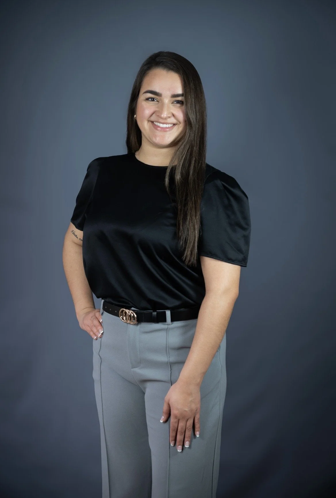A woman with long dark hair, smiling, wearing a black satin short-sleeve top, light gray pants, and a black belt with a gold buckle, standing against a plain dark background.