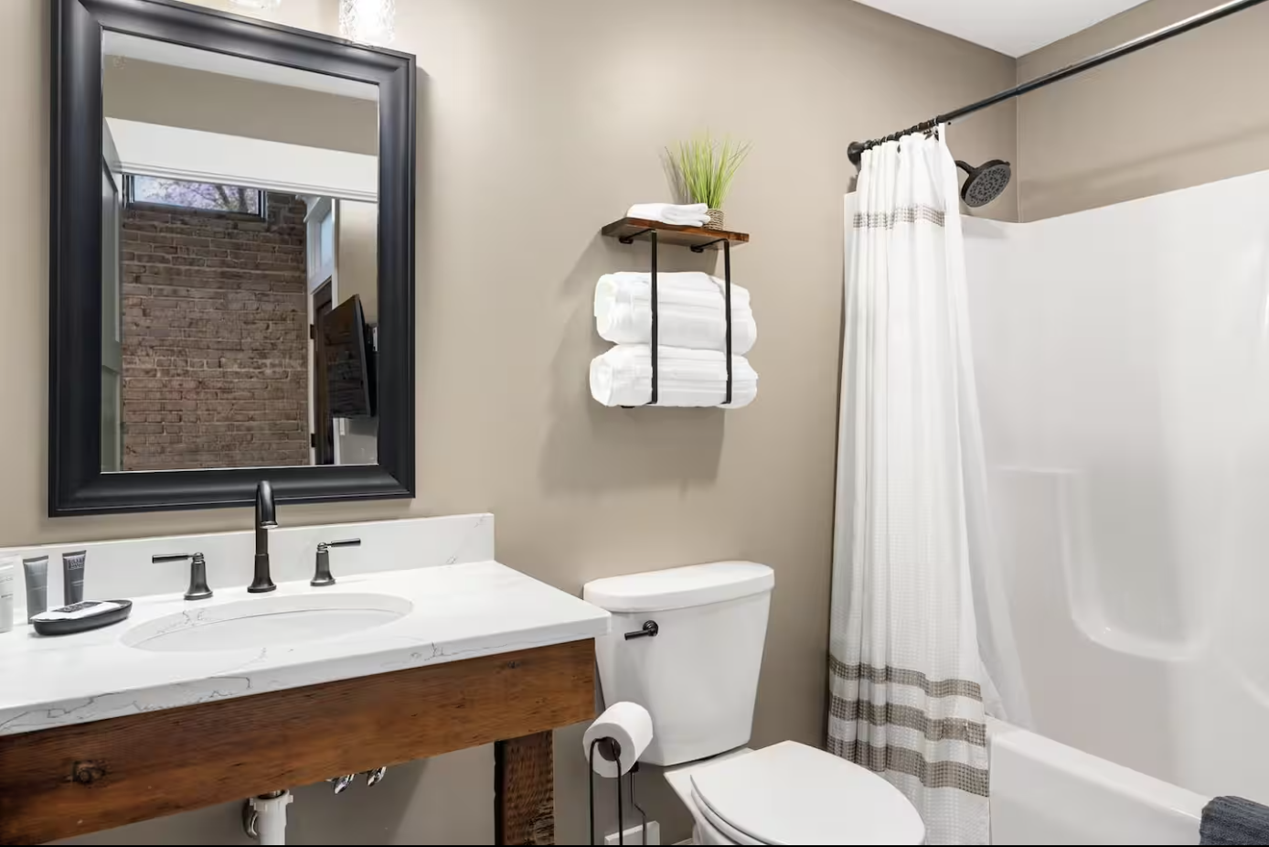 Bathroom with a white sink and black fixtures, a large mirror, a toilet with a roll of toilet paper, a wooden shelf with rolled white towels and a potted plant, a shower with a white curtain, and a brick wall reflection in the mirror.