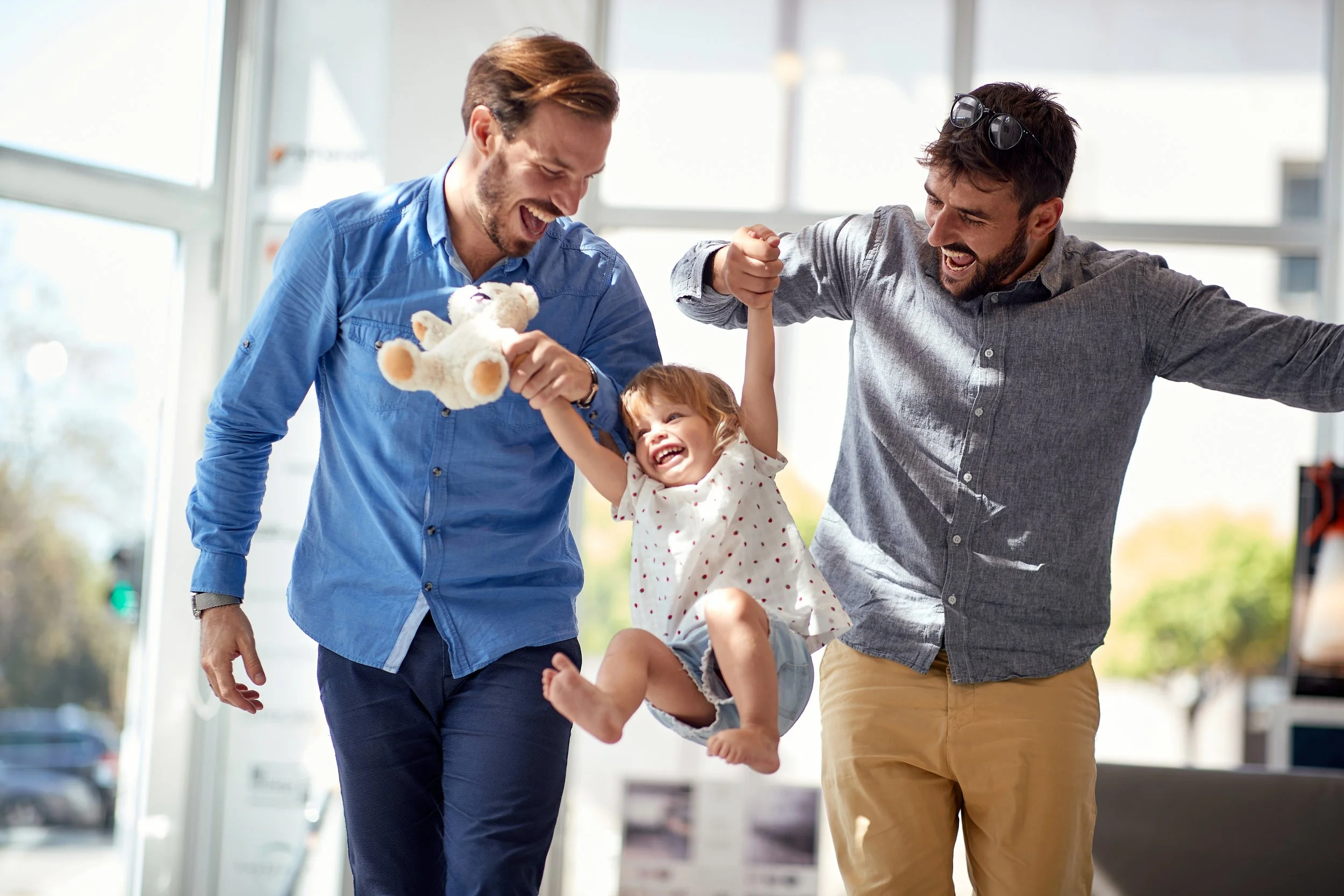 Two smiling adult men and a young girl bouncing and playing together indoors near large windows, with the girl holding a teddy bear.
