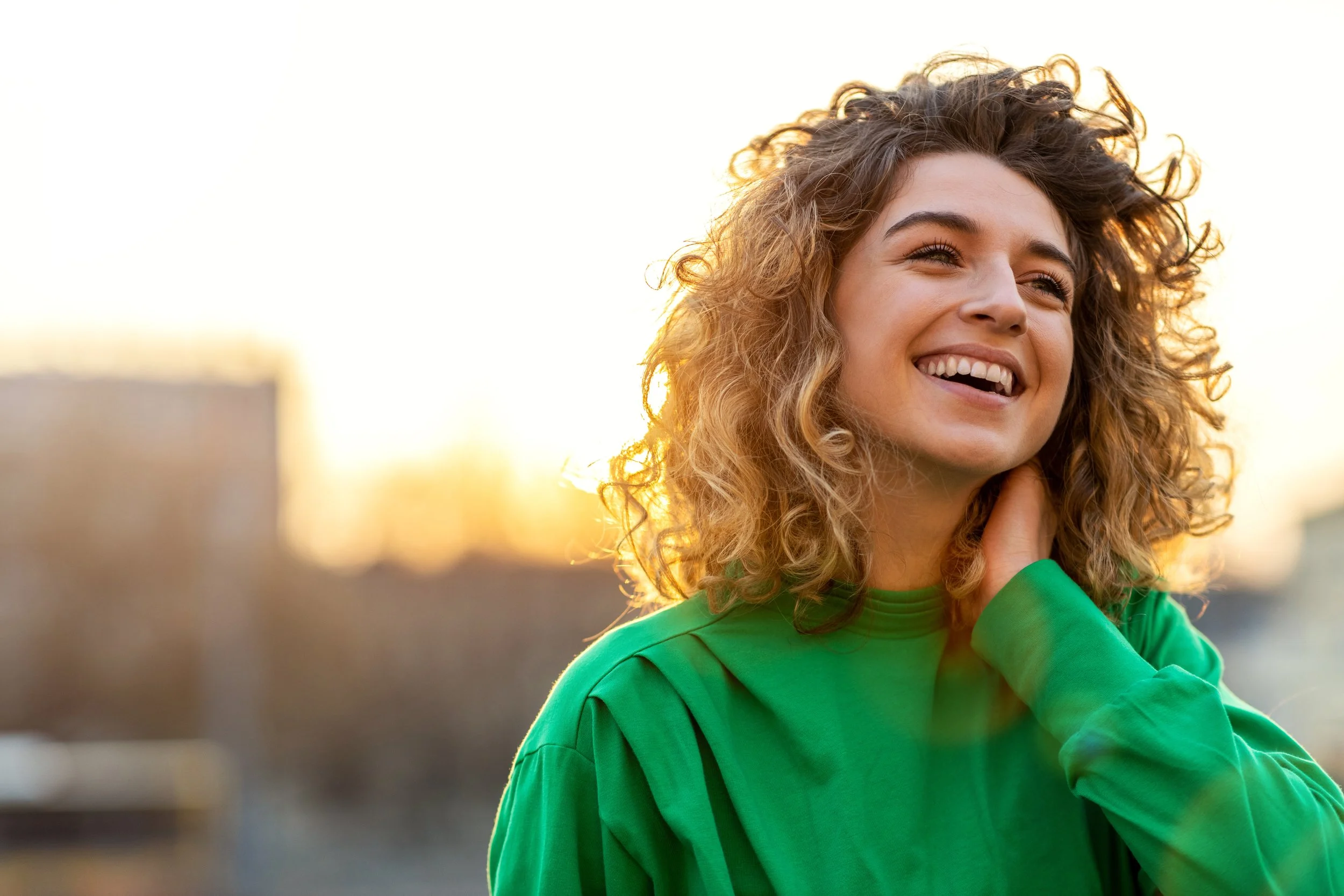 A young woman with curly hair wearing a green shirt, smiling and looking happy outdoors during sunset with blurred cityscape background.