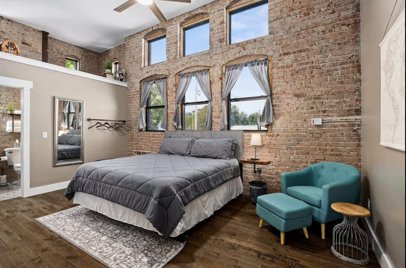A bedroom with exposed brick walls, hardwood floors, a bed with gray bedding, a teal armchair with an ottoman, a side table, and a lamp, with multiple windows and hanging curtains.
