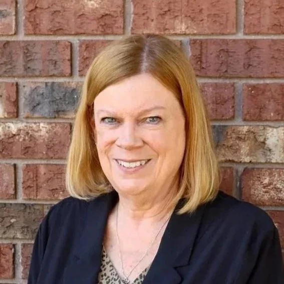 A woman with shoulder-length blonde hair smiling, wearing a navy blazer, colorful patterned top, and a gold necklace, standing in front of a brick wall.