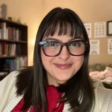 A woman with long dark hair, glasses, and a beige sweater smiling in a room with bookshelves and framed pictures on the wall.