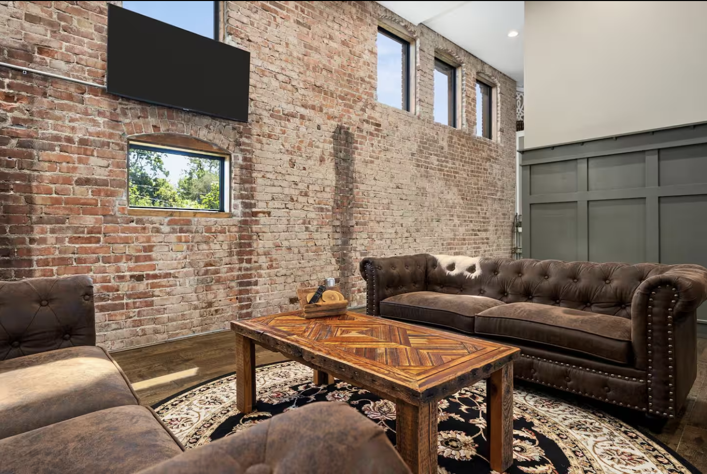 A cozy, industrial-style lounge with exposed brick walls, three small windows, dark leather sofas, a wooden coffee table, and a patterned rug.