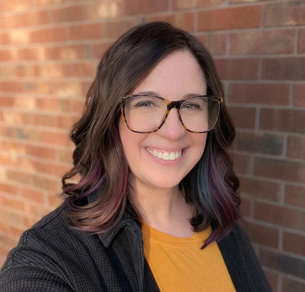 A smiling woman with glasses and wavy brown hair standing in front of a brick wall.