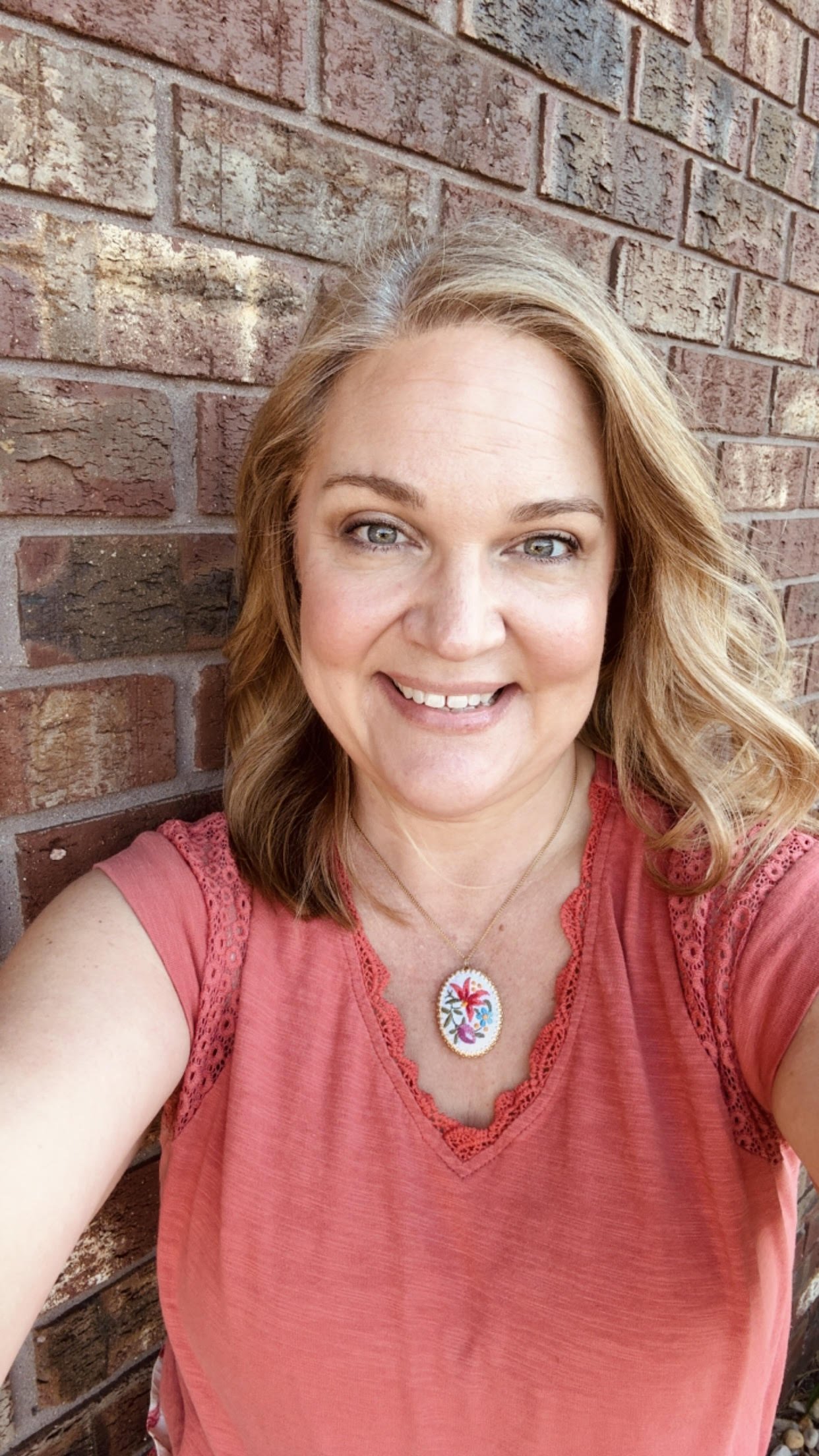 A woman smiling with blonde wavy hair, wearing a pink sleeveless top with lace details, a necklace with a colorful floral pendant, standing in front of a brick wall.