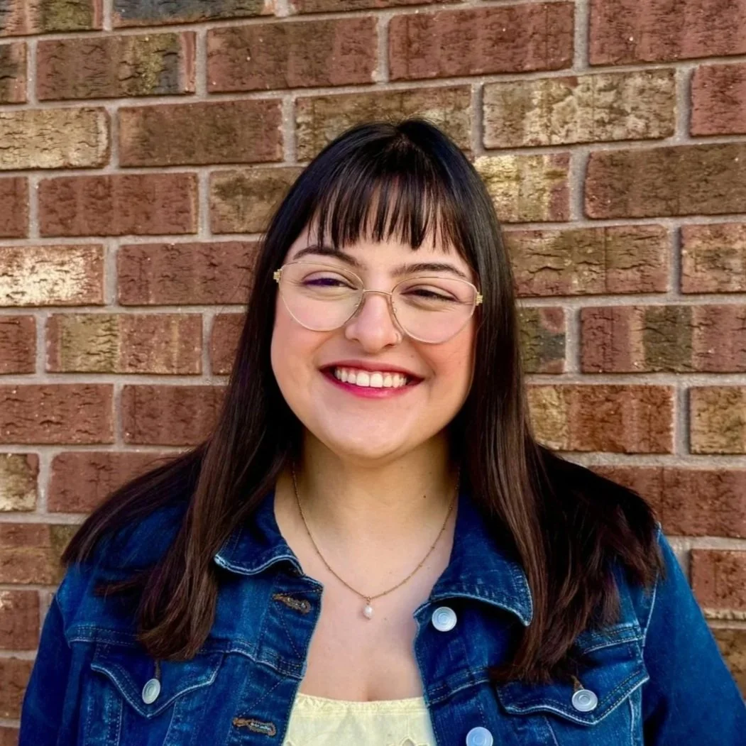 A woman with long dark hair, glasses, and a beige sweater smiling in a room with bookshelves and framed pictures on the wall.