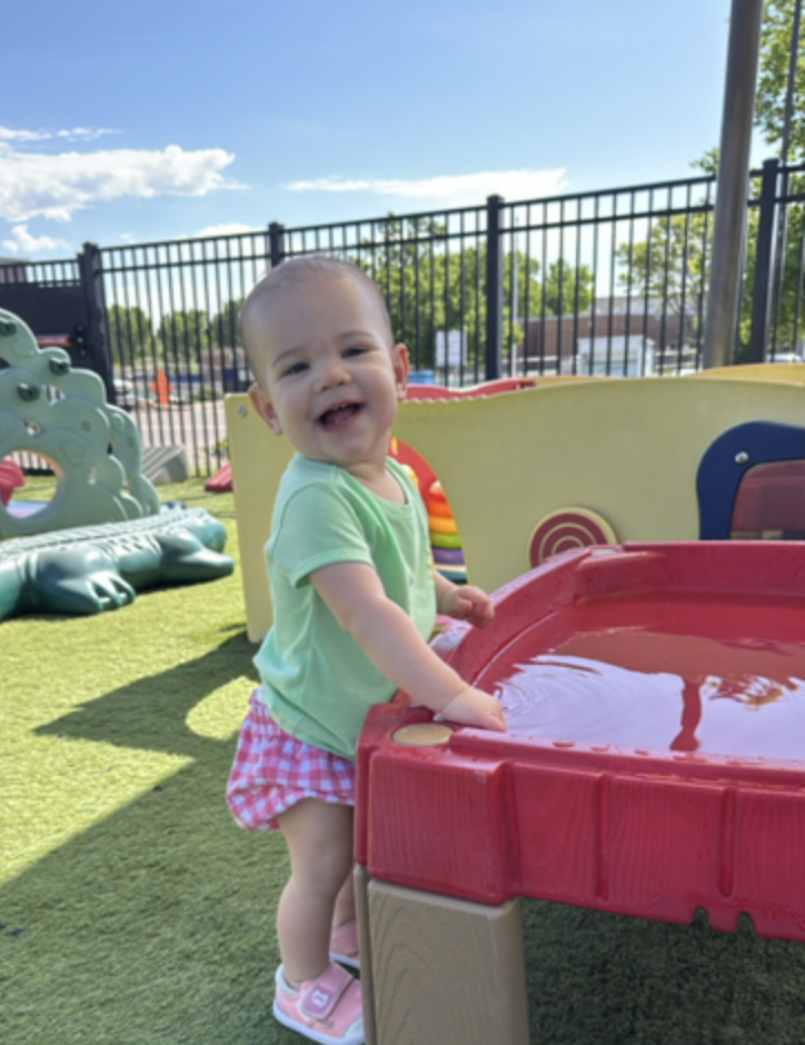 A young child playing with a water table outdoors on a sunny day, surrounded by playground equipment and a black fence.