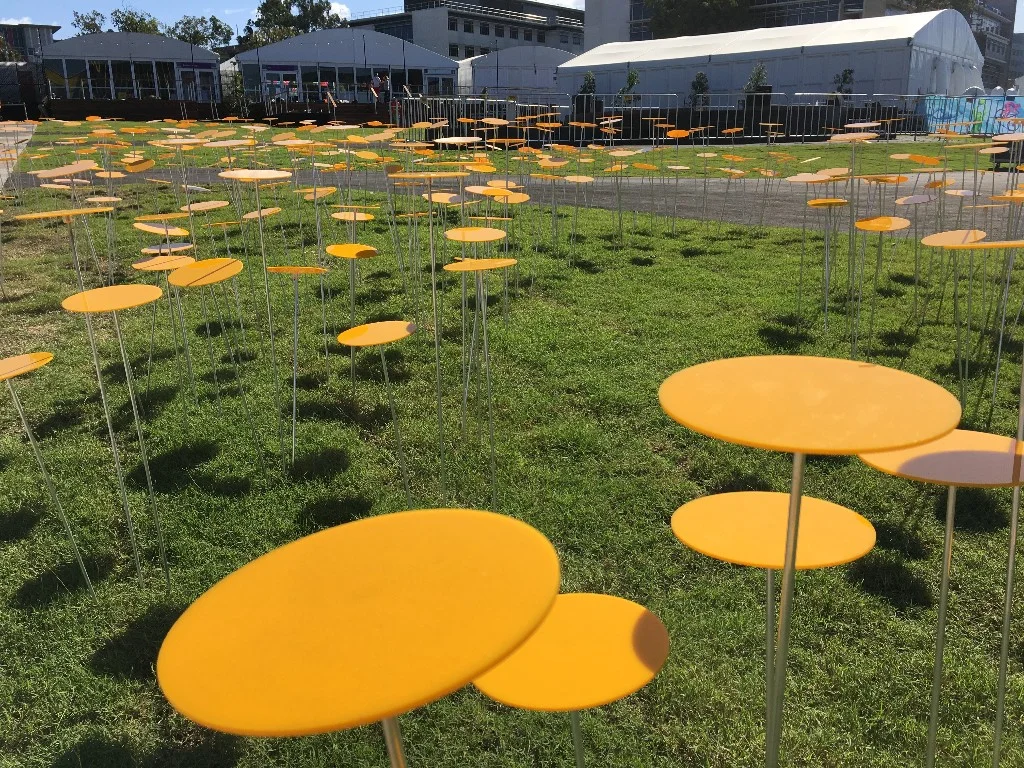 In 2006, Queensland artist Elizabeth Poole exhibited Blooming Yellow Wave for SWELL Sculpture Festival. The blooms stood gloriously amongst the Currumbin Beach dune, simulating the way wildflowers grow. For the Gold Coast 2018 Commonwealth Games, SWELL commissioned Elizabeth to rework the installation on a grand scale with the 853 medal winning athletes at its core as the Medal Blooms installation. Each solitary flower stands tall yet unified. Every bloom represents a multitude of stories, growing with the achievements and triumphs of the Games. As an artist, Elizabeth Poole plays at the intersections of art and nature. The Medal Blooms work responds directly to the surrounding local environment and draws upon the cheerful Climbing Guinea Flower, indigenous to the coastal regions of Eastern Australia. The climbing flower provides a metaphor for the Athletes competing at the Games, who are undertaking a climb of their own - to reach their personal peak at the top of their chosen field, being recognised by the highest awards of Gold, Silver and Bronze. The enthusiasm of colour and curvilinear energy rippling from a centre point invites the viewer to immerse themselves in the incredible achievements and feats unfolding during the Gold Coast 2018 Commonwealth Games as each bloom transforms with the medal winners names over the duration of the Games. VIEW THE MEDAL BLOOMS GALLERY >>>