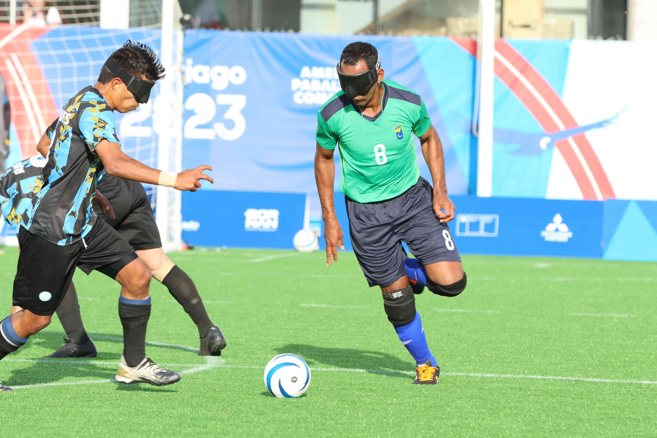 Two opposing blind soccer players run up towards the soccer ball from different directions.