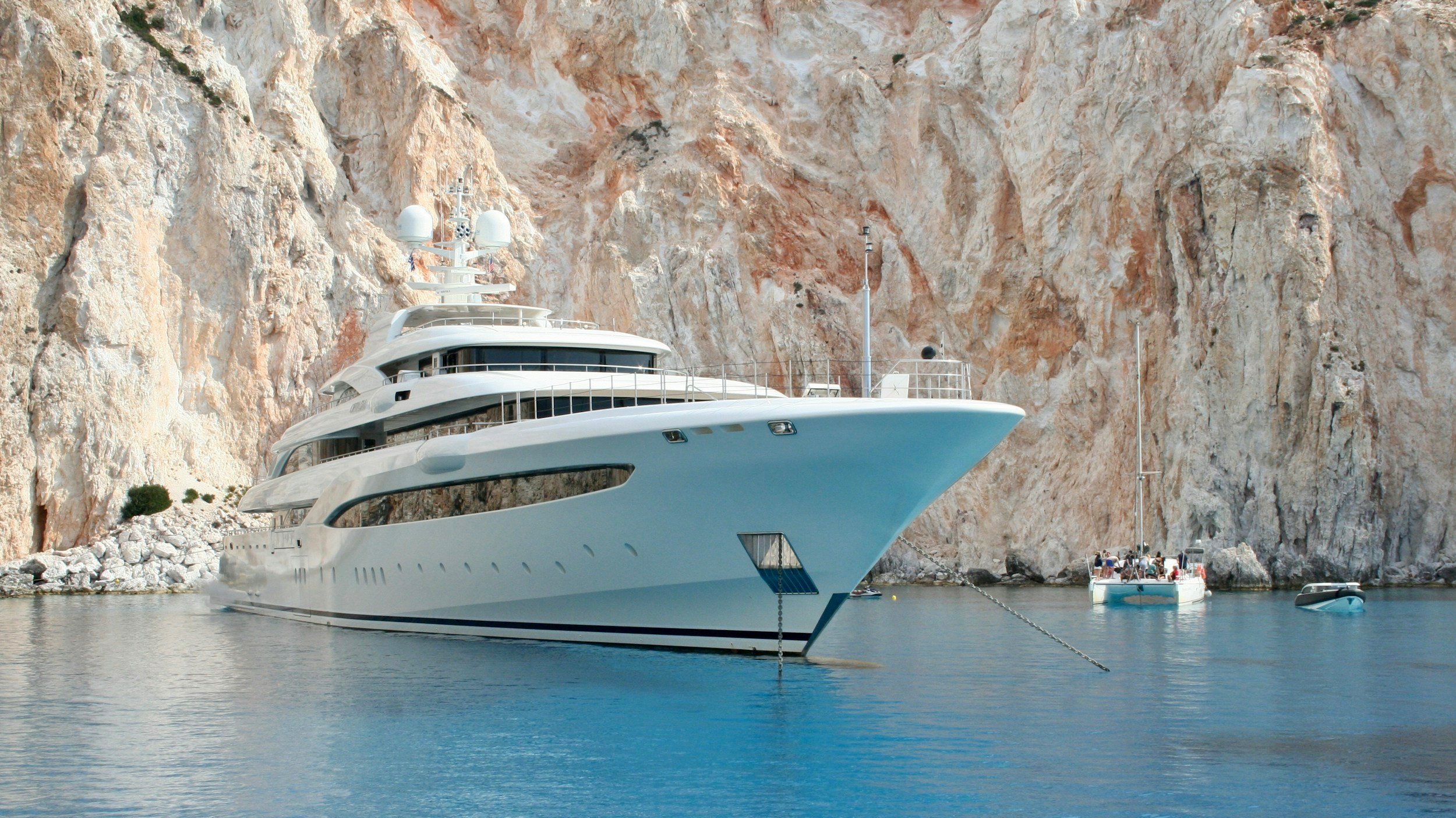A large white luxury yacht docked near a rocky, cliffside shoreline with several smaller boats nearby.