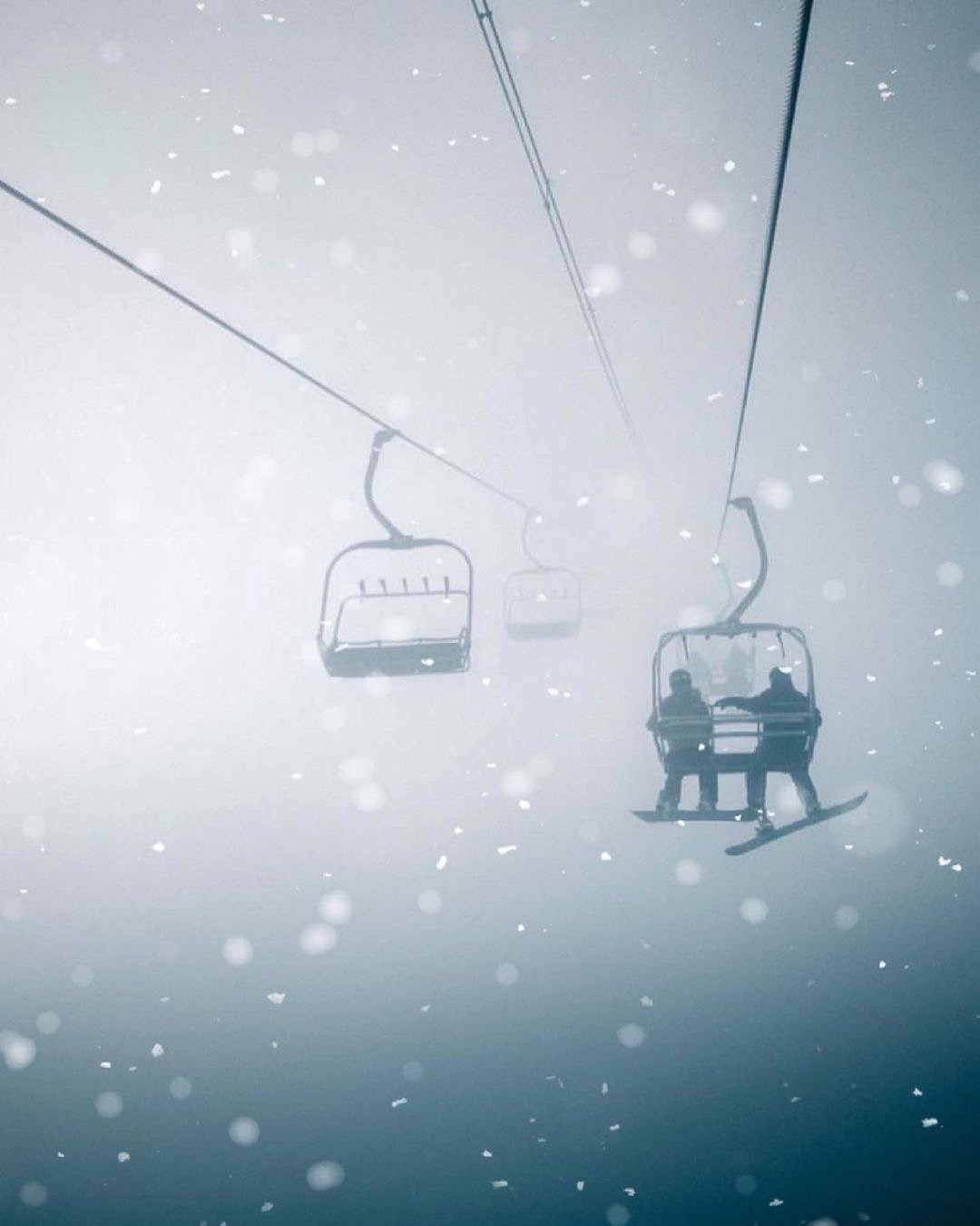 Two people riding a chairlift on a snowy day with falling snow and a foggy background.