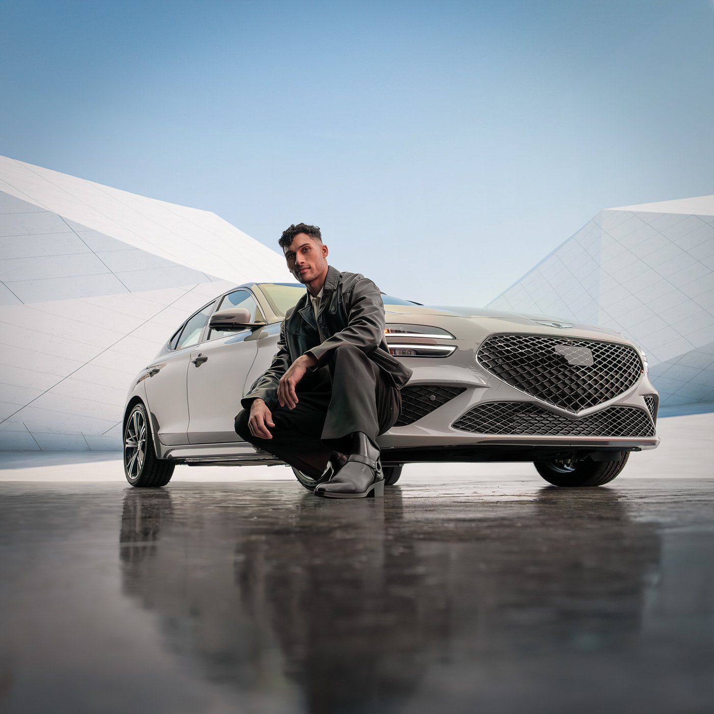 A man in dark clothing and gray boots crouches in front of a silver car with modern architecture in the background.