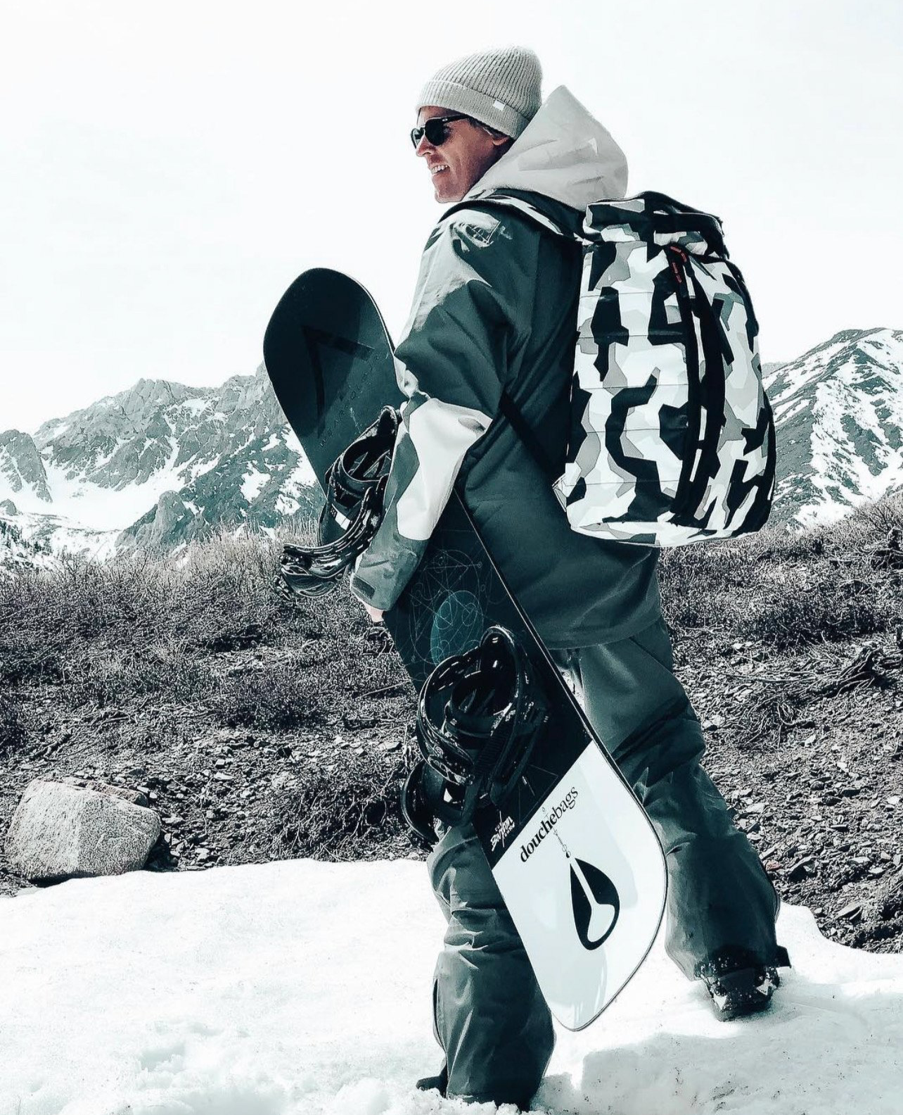 Man dressed in winter gear holding a snowboard in a snowy mountain landscape
