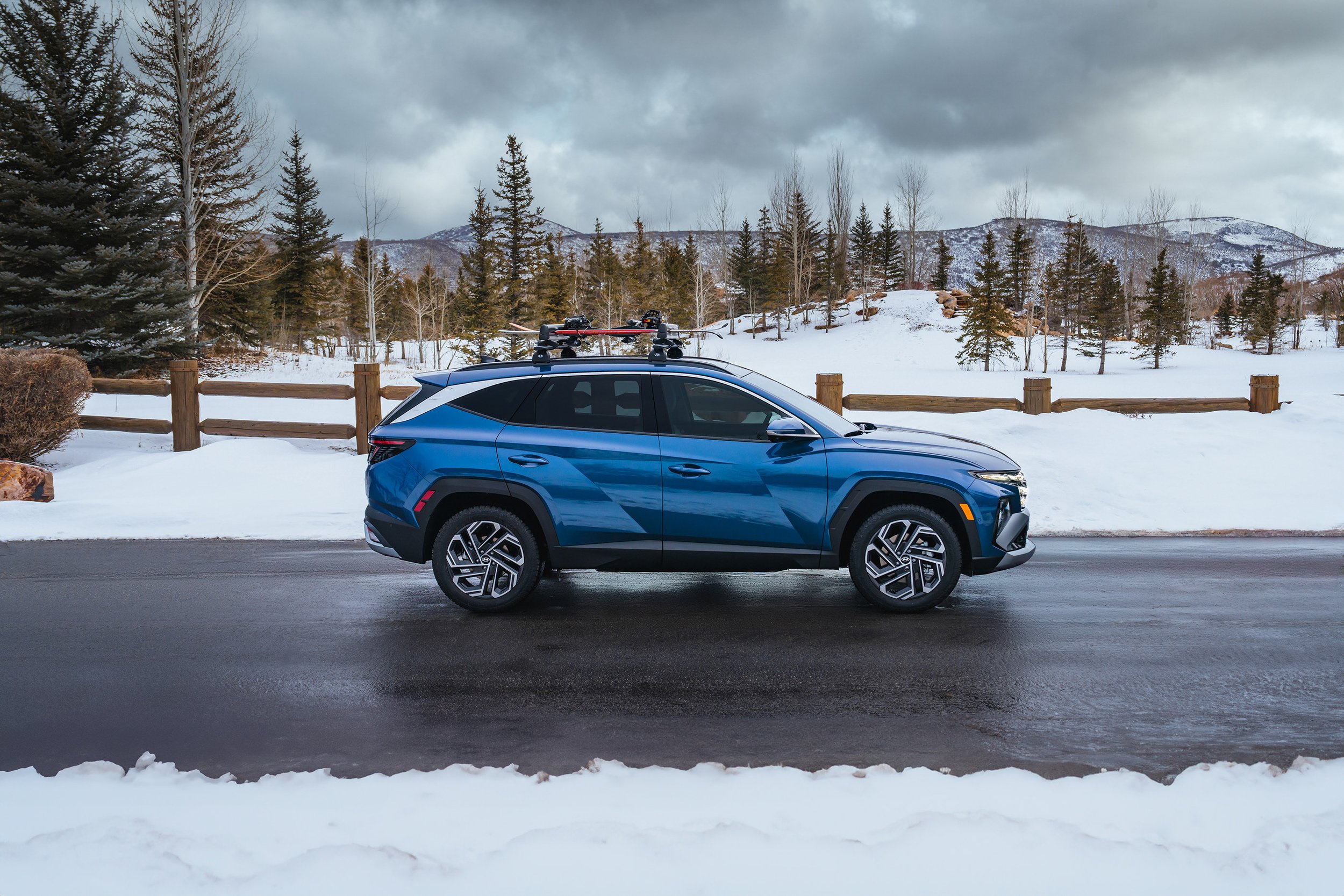 A blue SUV parked on a wet road with snow, trees, and mountains in the background on a cloudy day.