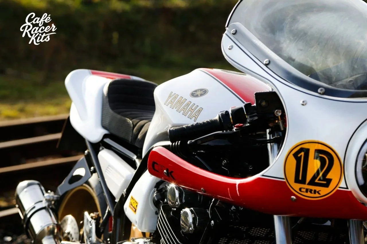 Close-up of a vintage Yamaha racing motorcycle with a white and red fairing, black seat, and a yellow racing number 12 on it, near train tracks.