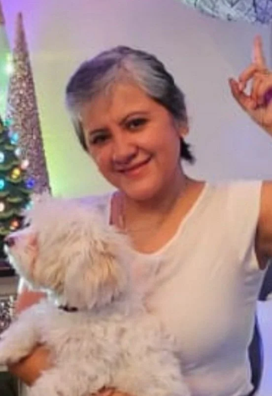Woman with short gray hair smiling and holding a fluffy white cat, standing next to a decorated Christmas tree with colorful ornaments and lights.