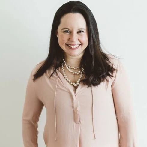A woman with long dark hair, wearing a light pink blouse and layered pearl necklaces, smiling against a plain light background.