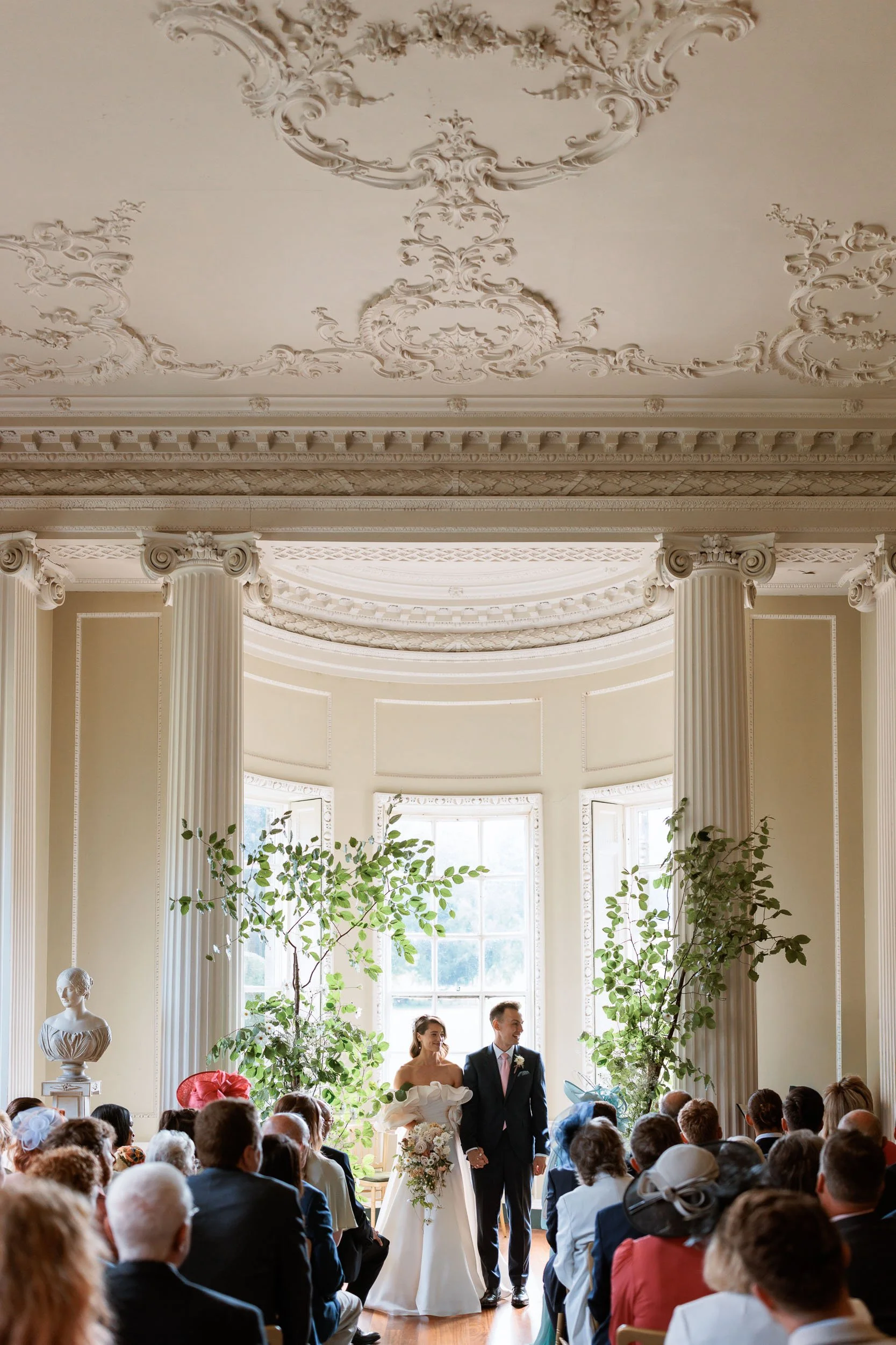 Bride and groom standing together during their wedding ceremony at Newburgh Priory, surrounded by guests beneath an ornate Georgian plasterwork ceiling.