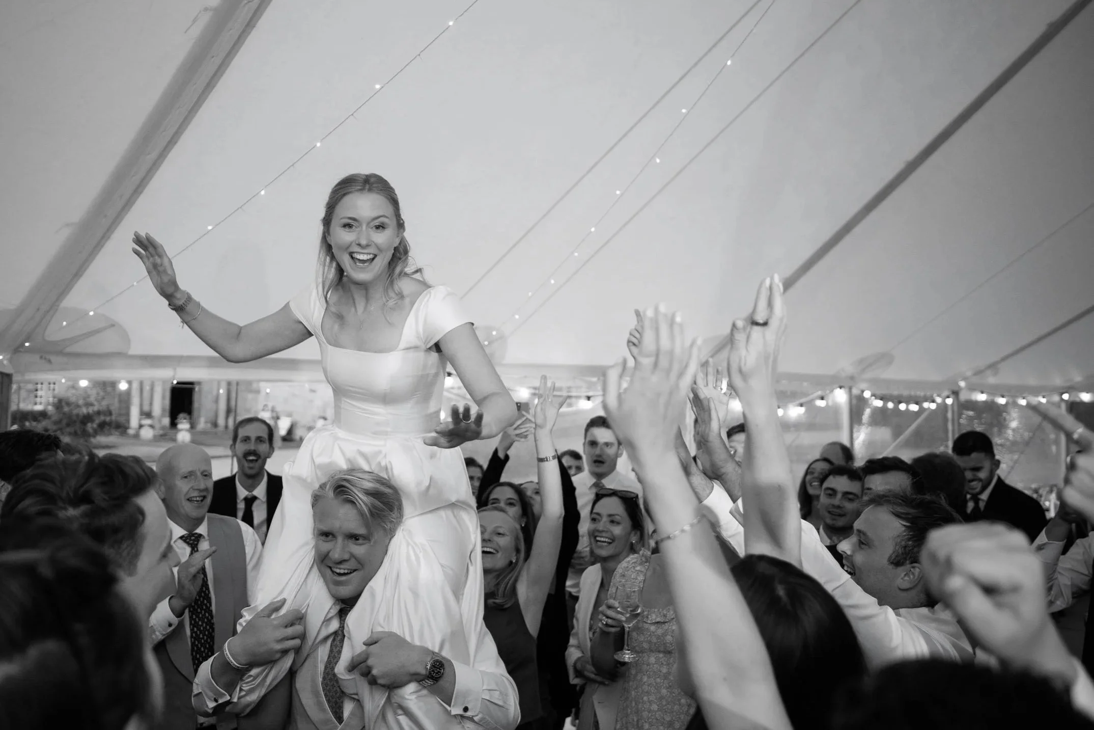 Black and white photograph of a bride being carried on someone's shoulders as wedding guests cheer during an evening celebration at Newburgh Priory.