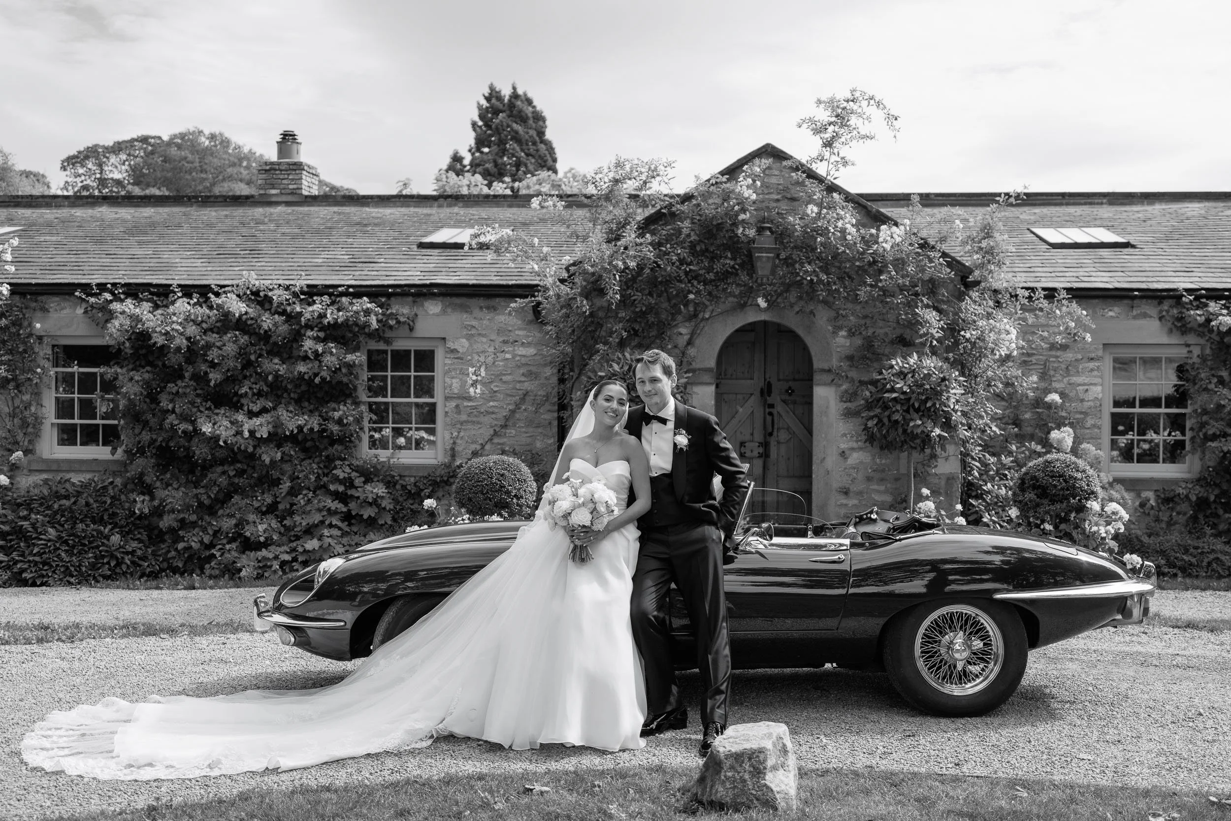 Black and white portrait of newlyweds beside a vintage wedding car at Middleton Lodge.