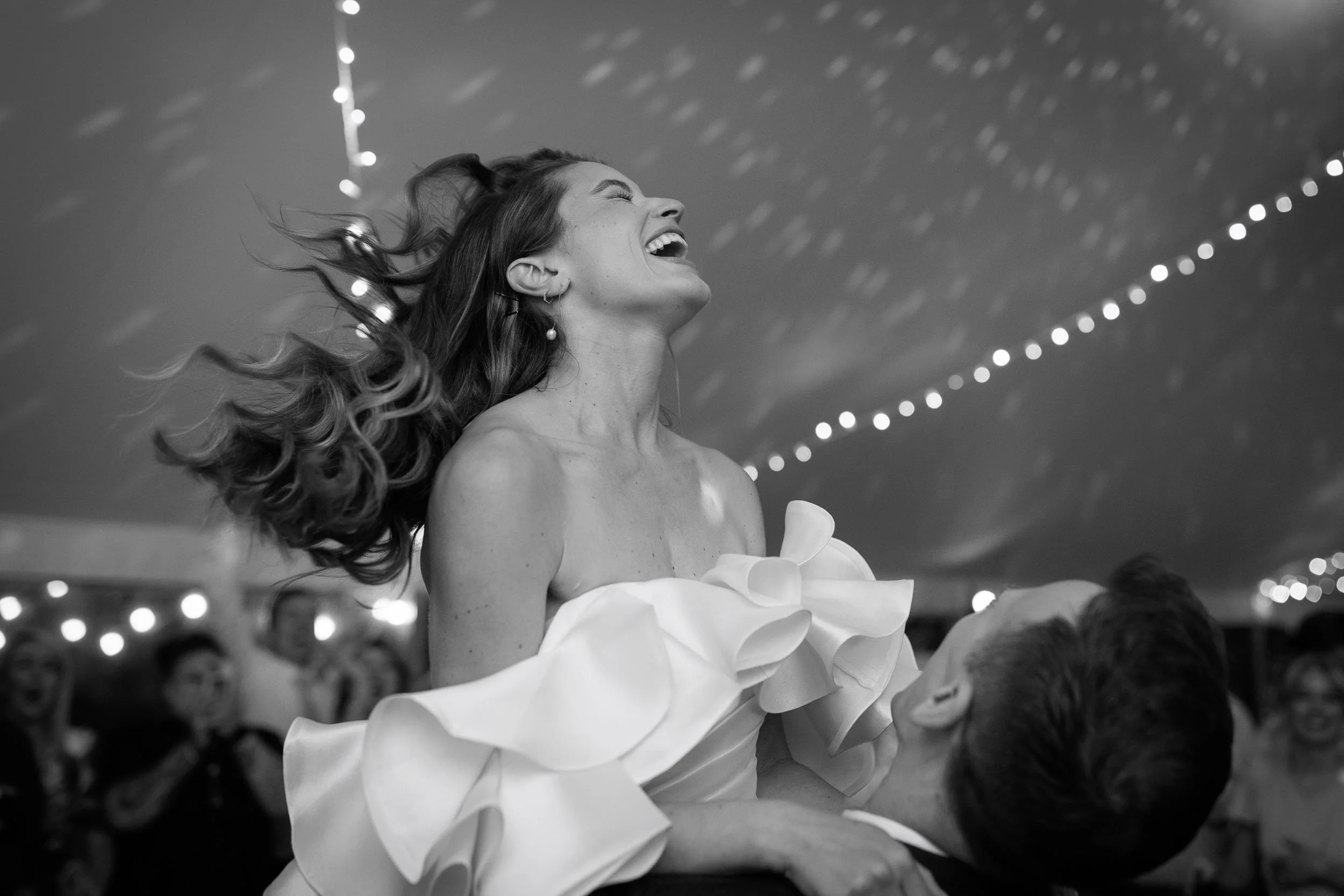 Black and white photograph of a bride laughing as she is lifted during the evening celebrations inside a wedding marquee at Newburgh Priory.