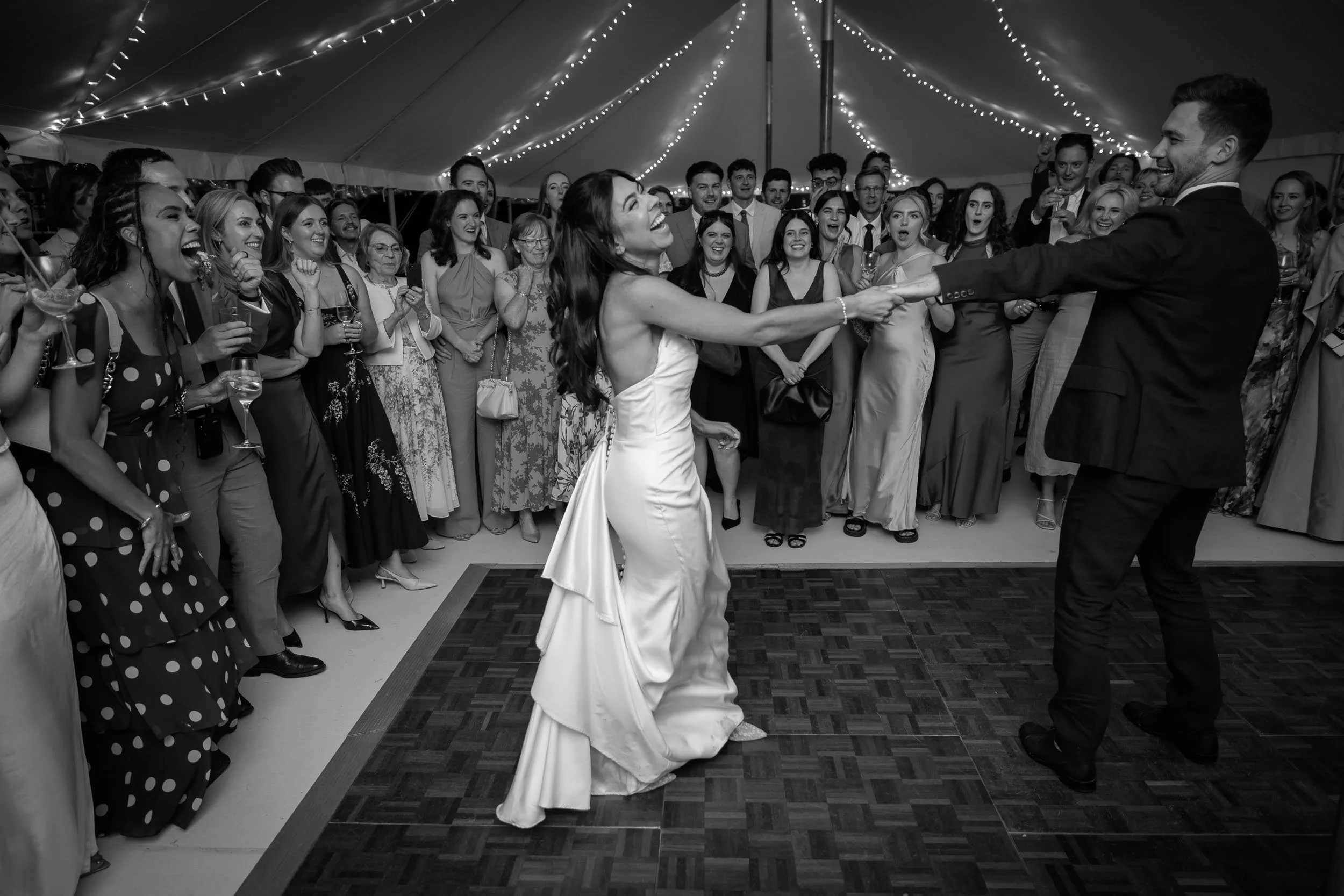 Bride and groom dancing together beneath festoon lights in a marquee at Newburgh Priory, surrounded by cheering wedding guests.