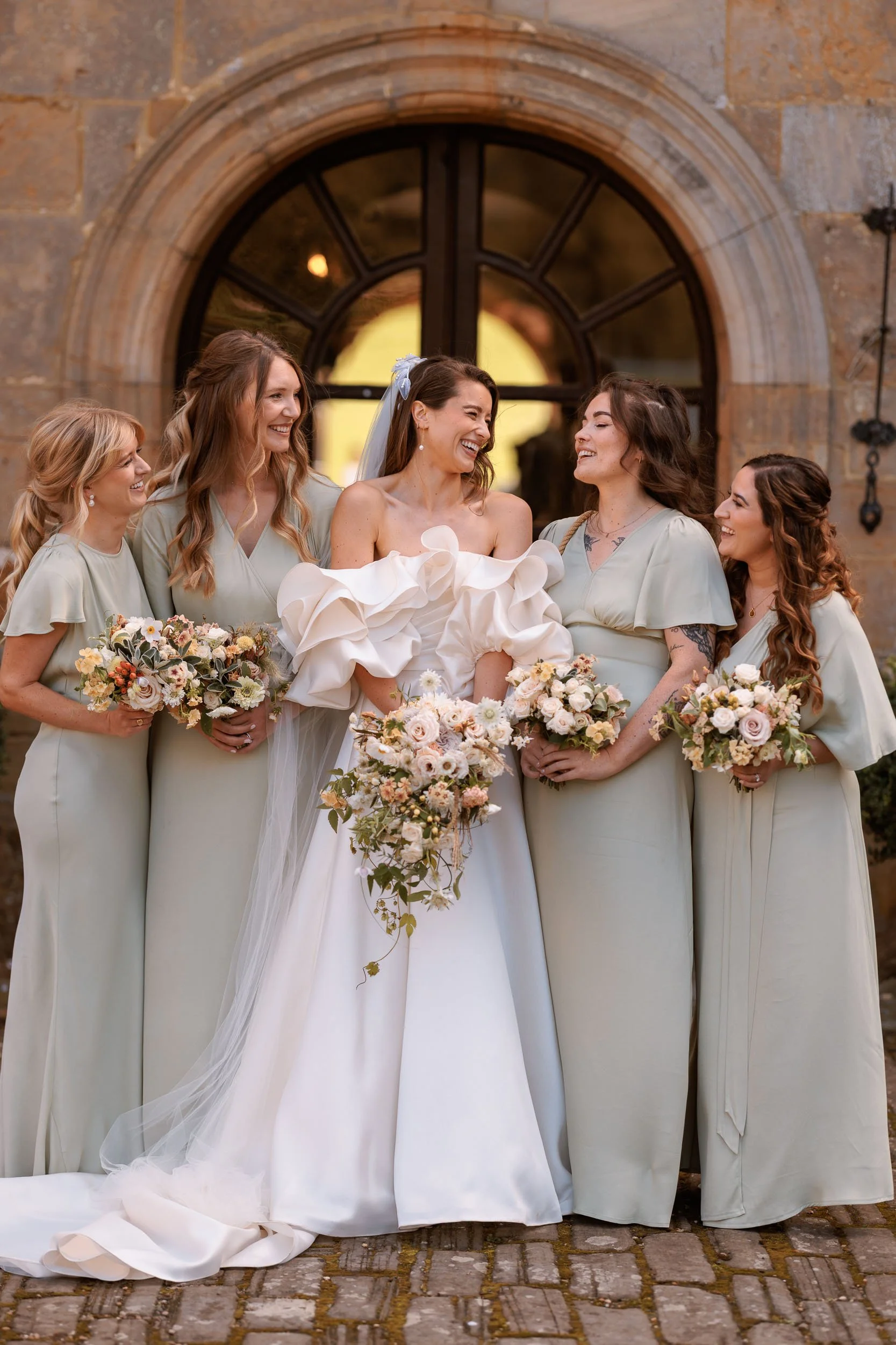 Bride laughing with her four bridesmaids outside a historic stone archway at Newburgh Priory in North Yorkshire.