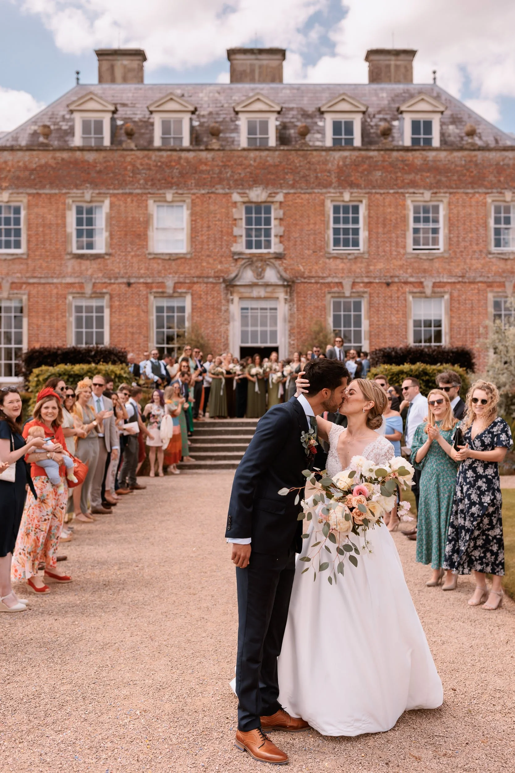 Newlyweds sharing a kiss outside St Giles House in Dorset, surrounded by wedding guests celebrating in front of the historic country house.