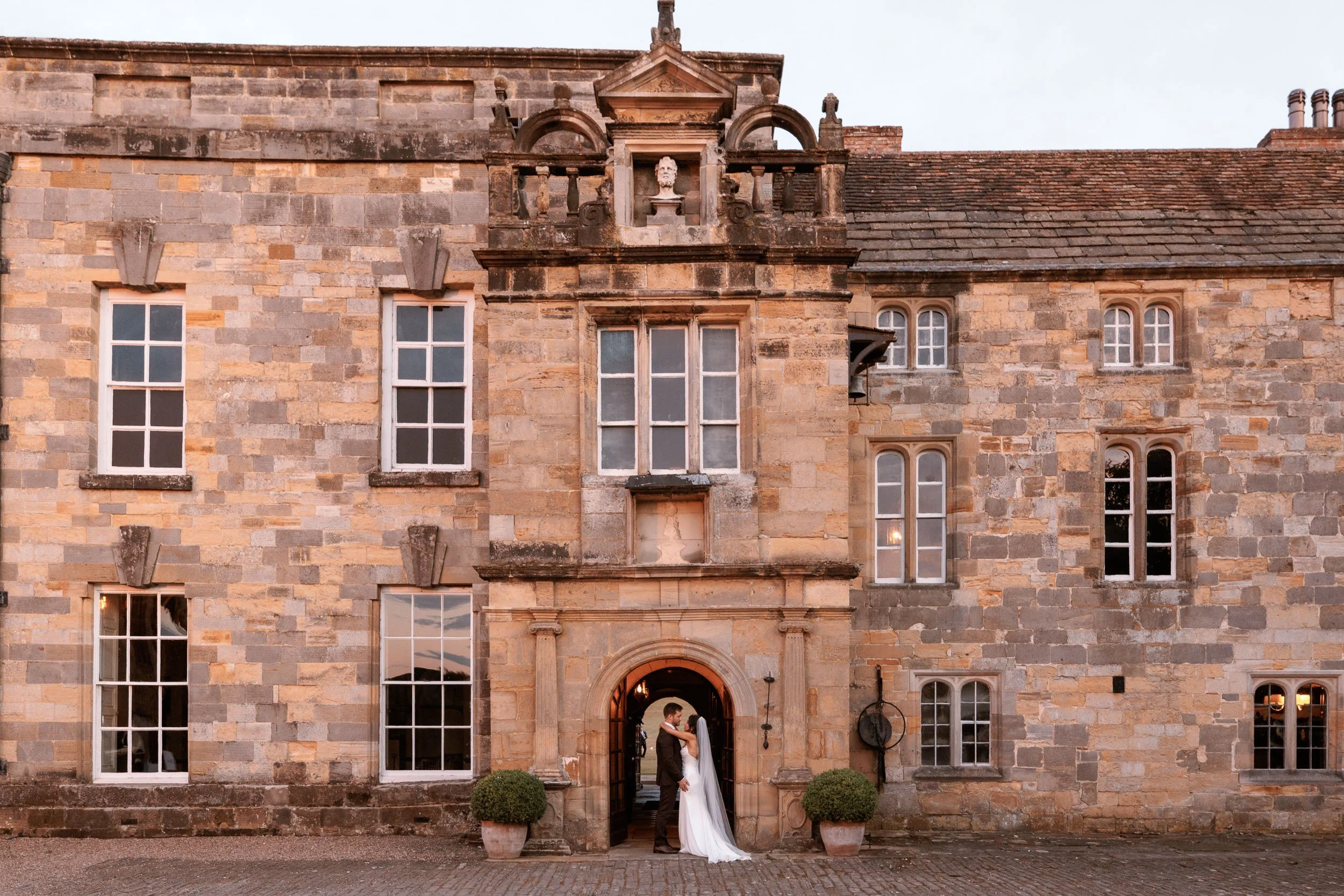 Bride and groom embracing beneath the historic stone entrance archway at Newburgh Priory, North Yorkshire.
