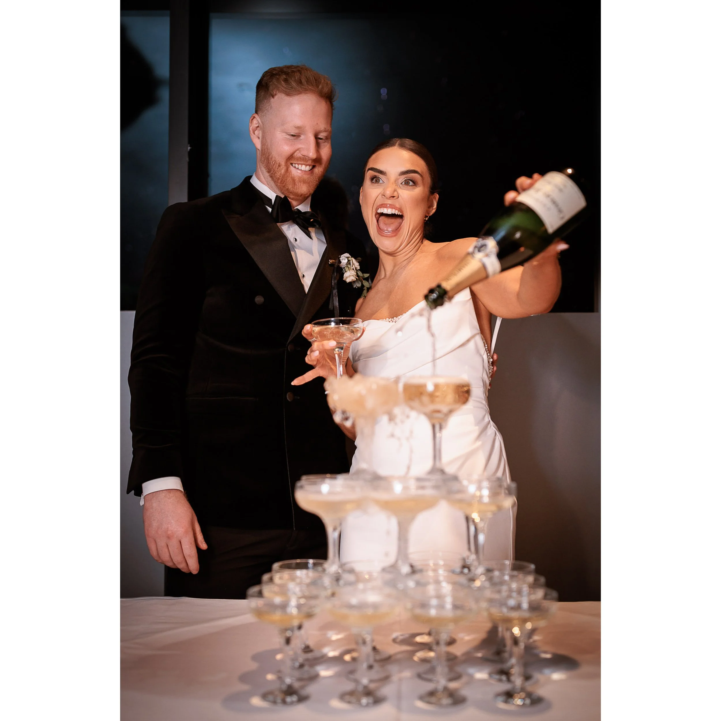 A couple in wedding attire pouring champagne into a tower of coupe glasses at an outdoor celebration.