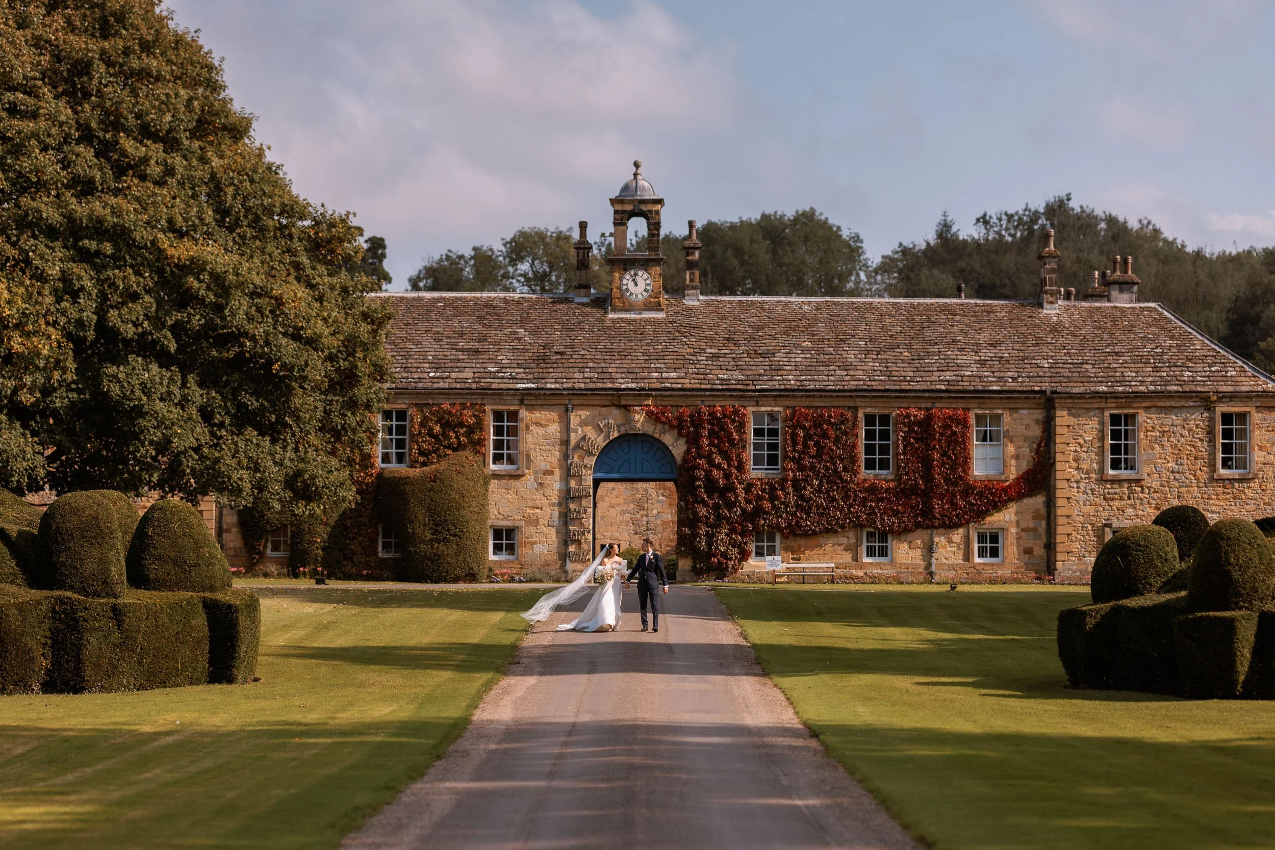Bride and groom walking along the drive at Newburgh Priory, with its historic stone clock-tower building and formal gardens behind them.