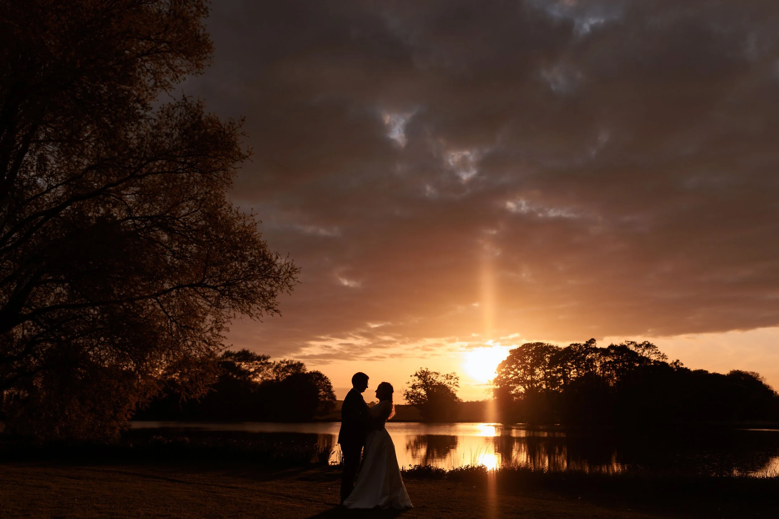 Silhouette of a bride and groom beside the lake at Newburgh Priory, with golden sunset light reflecting across the water.
