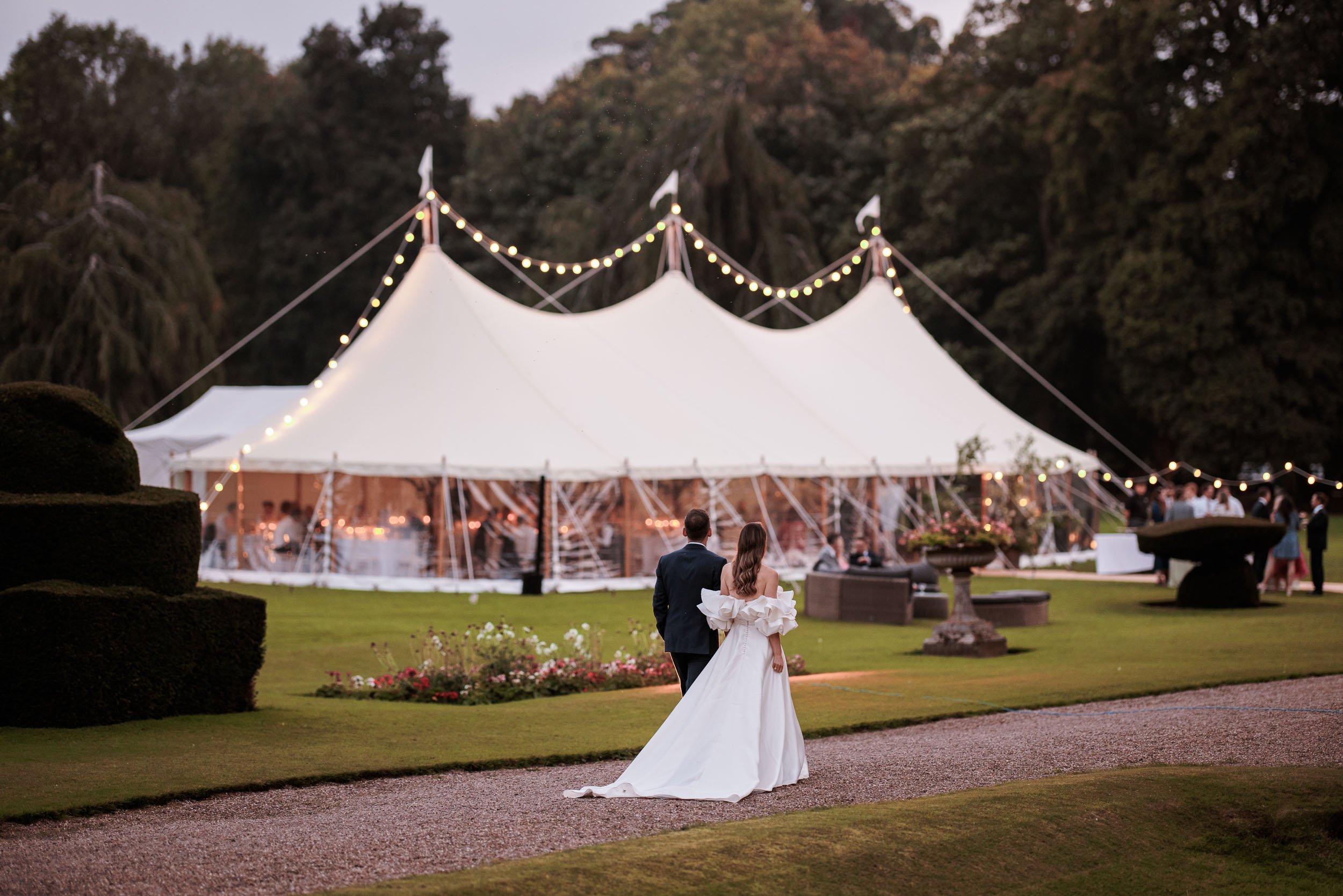 Bride and groom walking towards an illuminated wedding marquee at Newburgh Priory, with festoon lights glowing across the gardens at dusk.