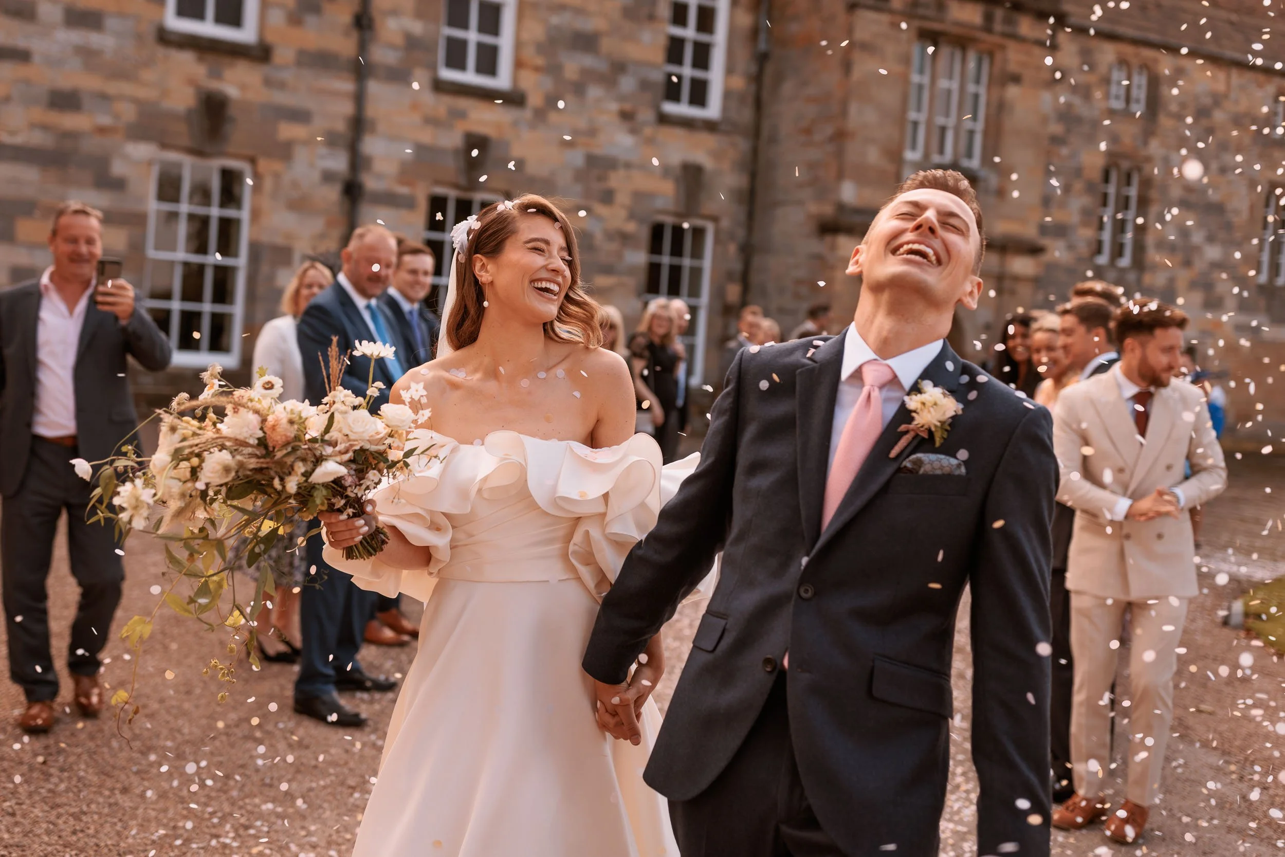 Newly married couple laughing as confetti falls around them outside Newburgh Priory, with wedding guests celebrating behind them.
