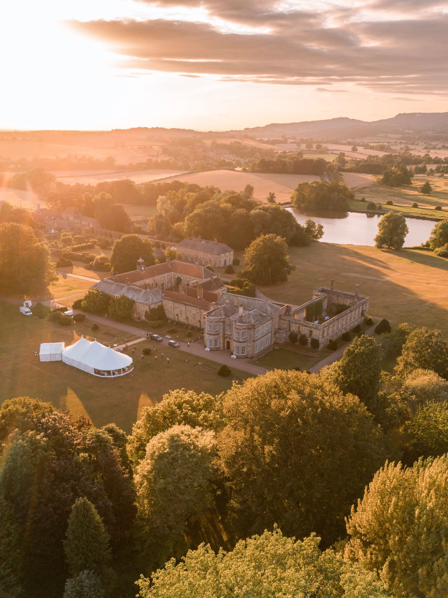 Aerial view of Newburgh Priory in North Yorkshire at golden hour, showing the historic buildings, ruins, wedding marquee and lake.