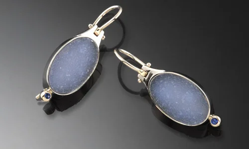   Oval hinge earrings in chalcedony druzy and 18K white gold. 2mm blue sapphire accents. Earrings snap shut securely at the back. Length 1.25" Width 0.5" Depth 0.25"     