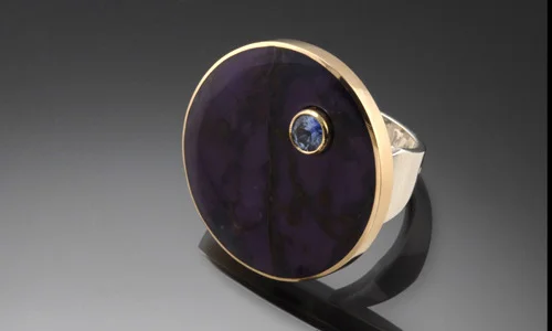   Inlay ring in 18K yellow gold, sterling silver, sugilite and 4.5mm faceted pastel sapphire accent. Diameter 1" Shank width 0.2" - 0.4" ﻿  