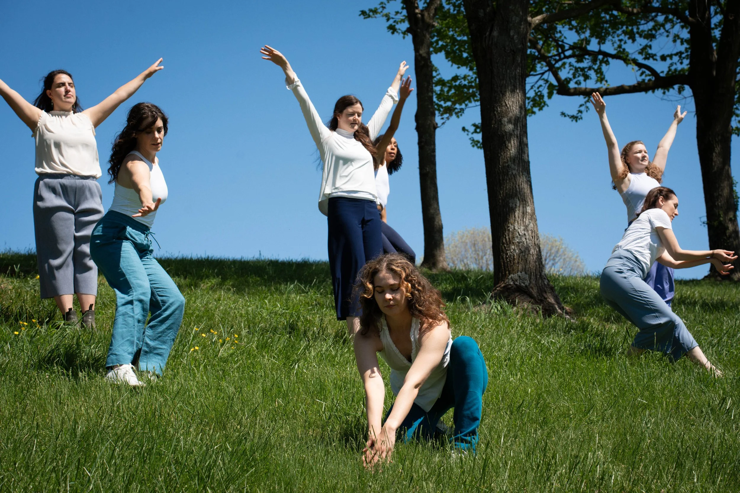 Six dancers in white tops and blue pants stand on a grassy hillside reaching their arms in different directions