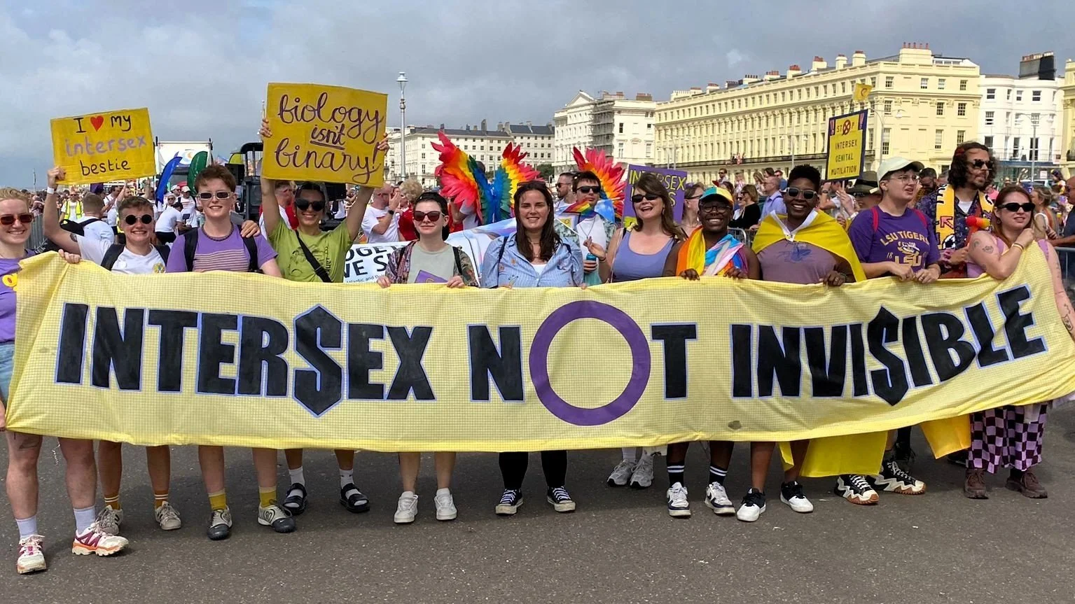  A diverse group of people at a Pride festival marching behind a banner that says 'Intersex not invisible'. 