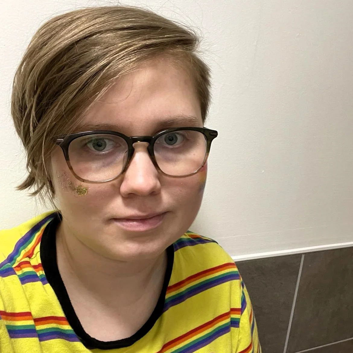  Beth getting ready for Pride, wearing glitter face paint and a rainbow striped tshirt. 