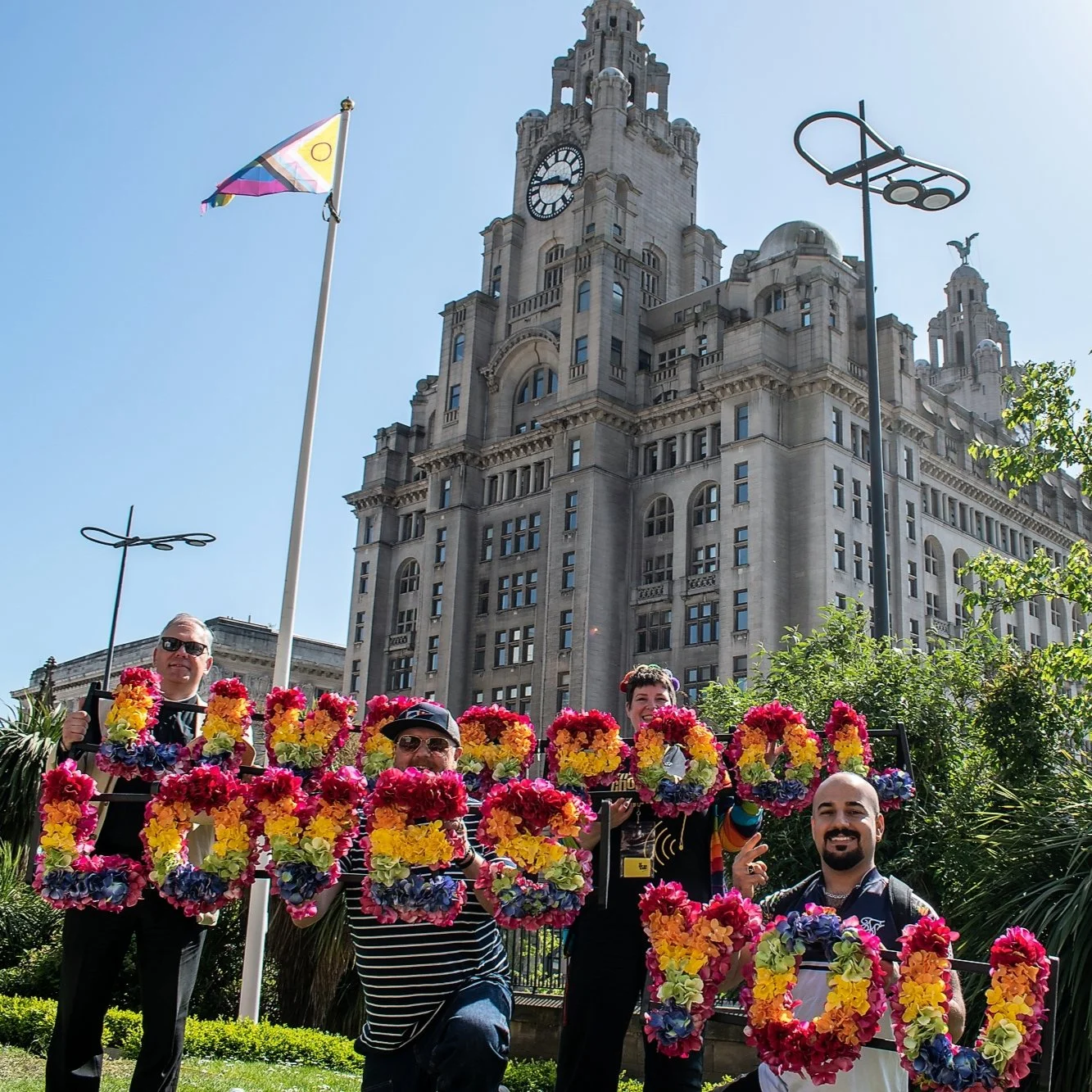 The Rector of Liverpool and friends hold flowers spelling out ‘Liverpool Loves You’ at the city’s parish church on Saturday 14th May, in front of the iconic Liver Building and a flagpole flying the Progress Pride flag . PHOTO: Rian Bailey-Weir