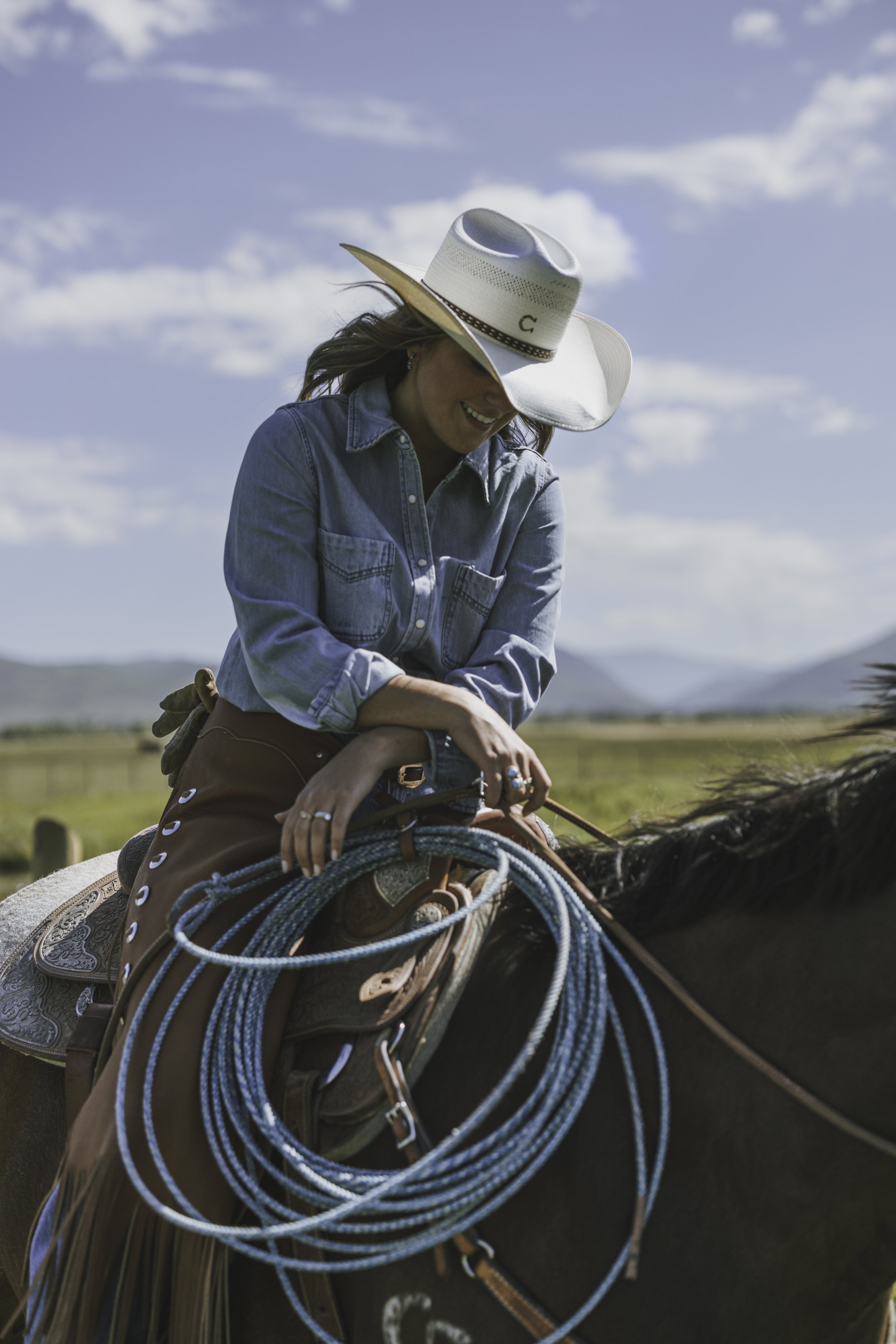 Cowgirl in denim and fringe sitting on horseback holding a coiled rope in an open ranch landscape.