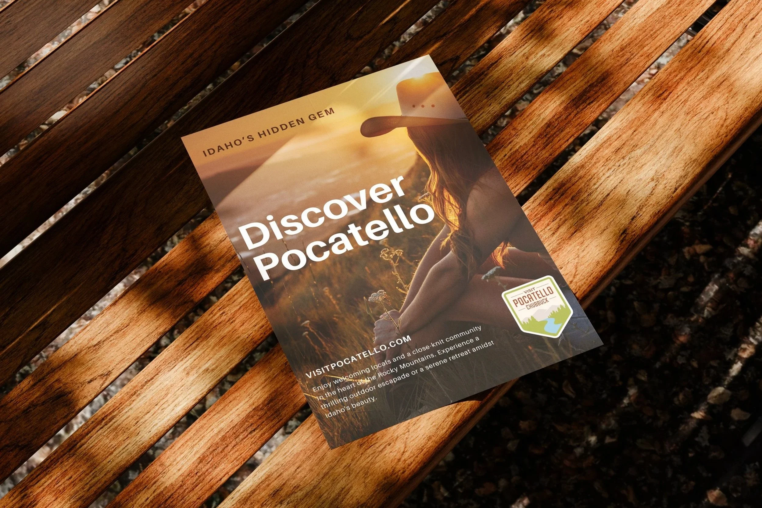 A brochure titled 'Discover Pocatello' resting on a wooden bench. The brochure features an image of a woman wearing a hat, sitting outdoors during sunset in a natural setting, with the Idaho's hidden gem logo.