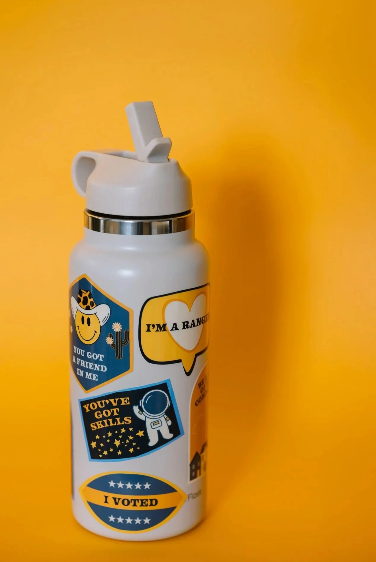 A white water bottle decorated with various stickers, including a smiley face wearing a hat, a space-themed sticker, a speech bubble that says 'I'm a Ranger,' a sticker that says 'You've Got Skills,' and a sticker that says 'I Voted,' set against a y