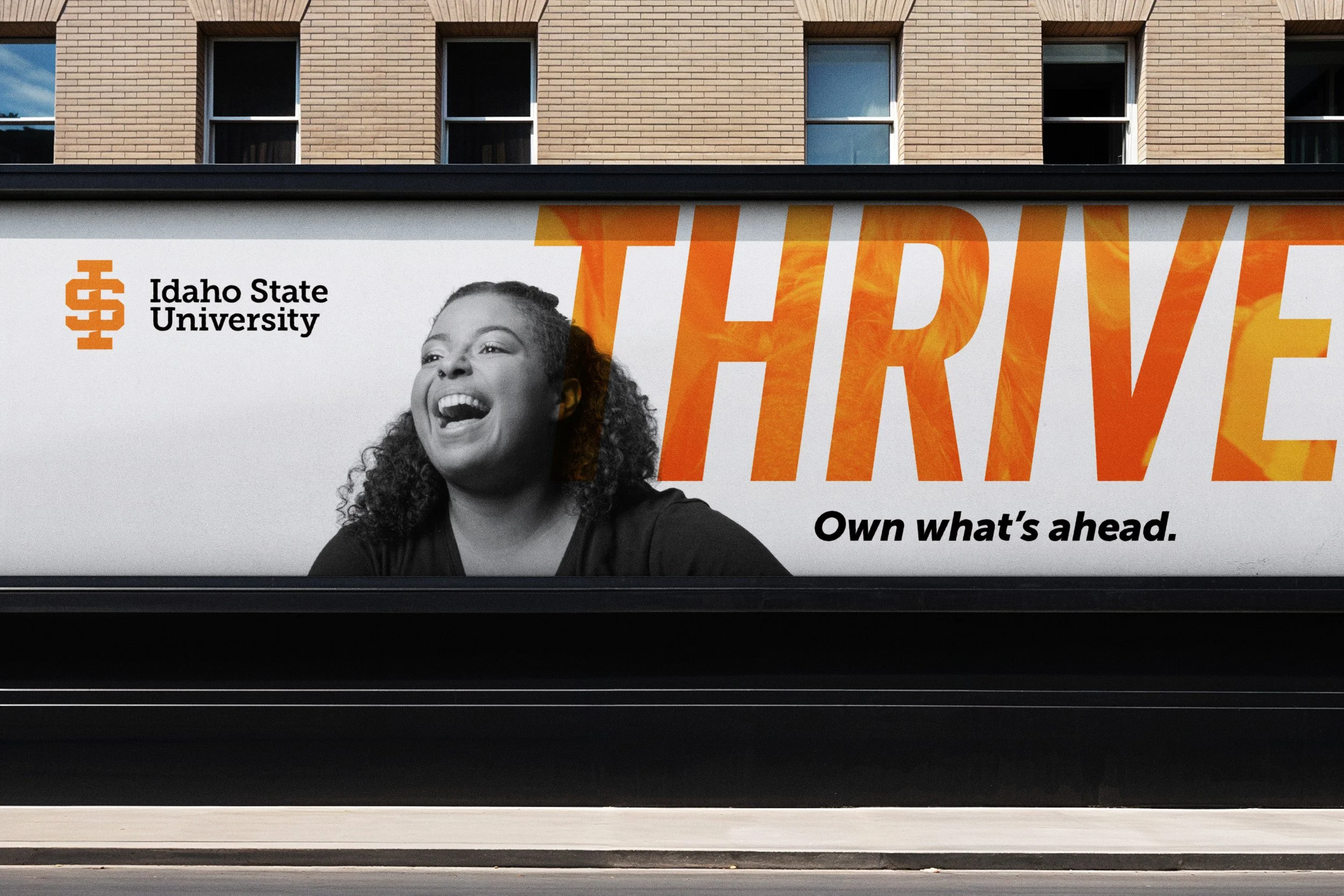 Billboard with Idaho State University logo, a black and white photo of a smiling woman, large orange text saying "THRIVE," and smaller black text saying "Own what's ahead." in front of a brick building.