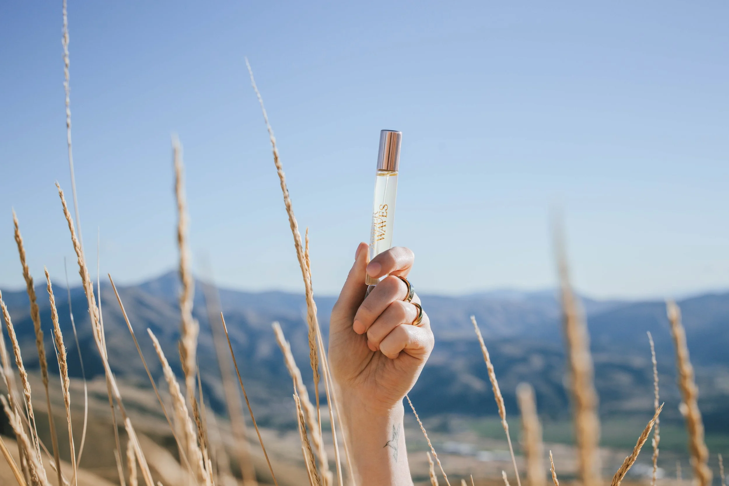 Hand holding Tru Western Waves purse spray fragrance against blue sky and mountain landscape in natural outdoor light.