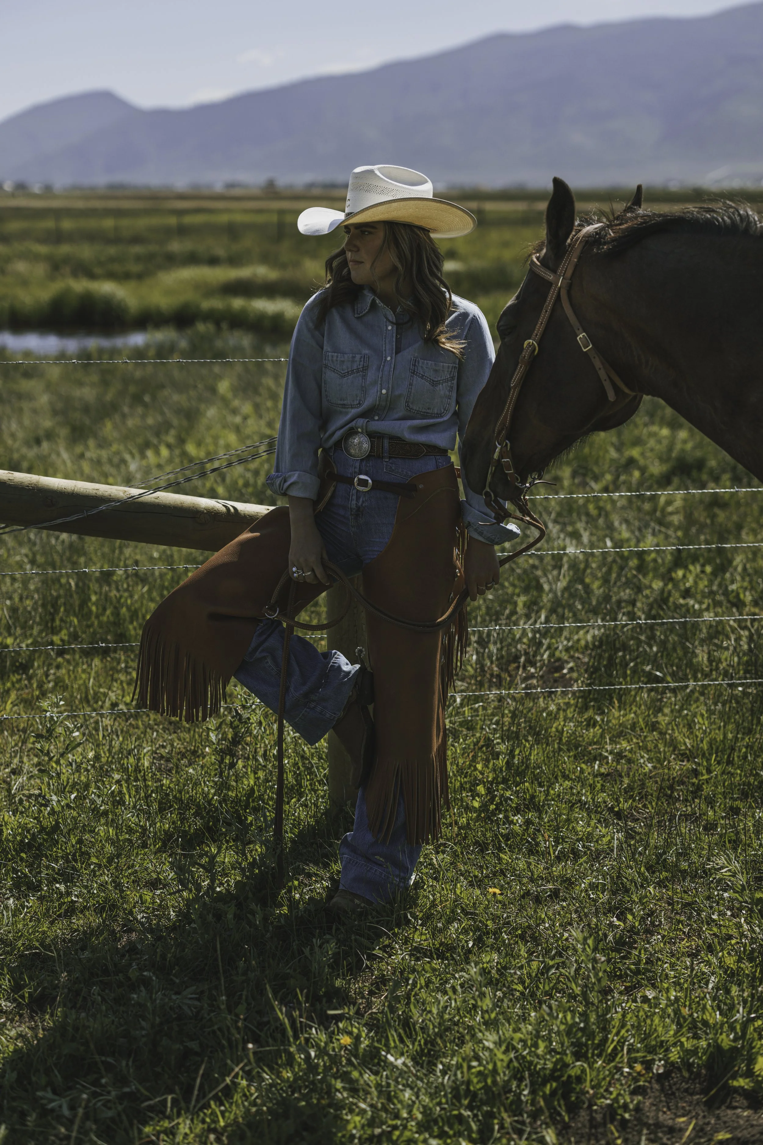 Cowgirl in a straw hat standing beside her horse in a sunlit pasture photographed for a western brand campaign.